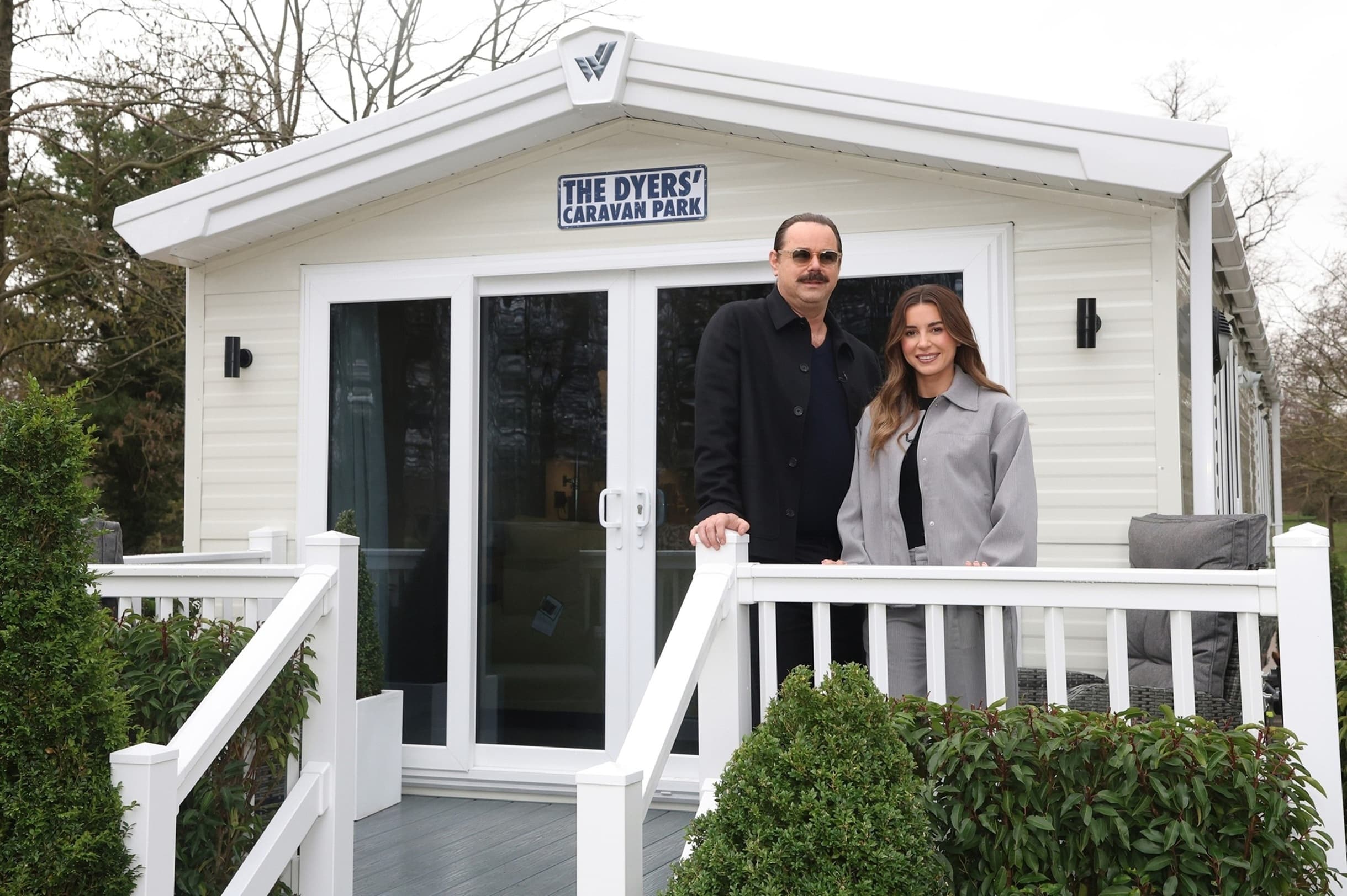 A man and woman stand outside a modern holiday home at The Dyers’ Caravan Park, surrounded by greenery.