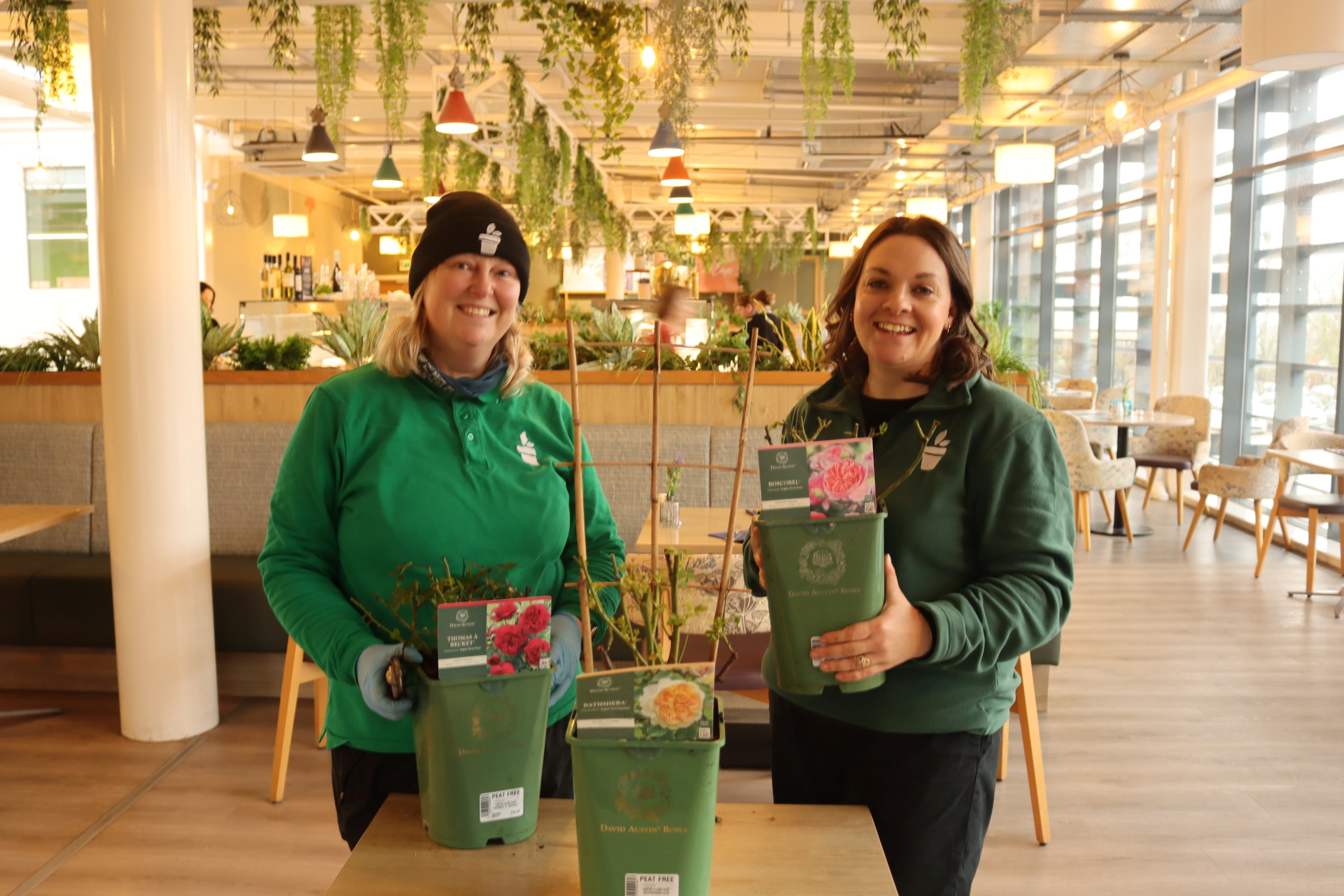 Two women in green attire hold potted rose plants inside a bright, modern garden center with hanging greenery.