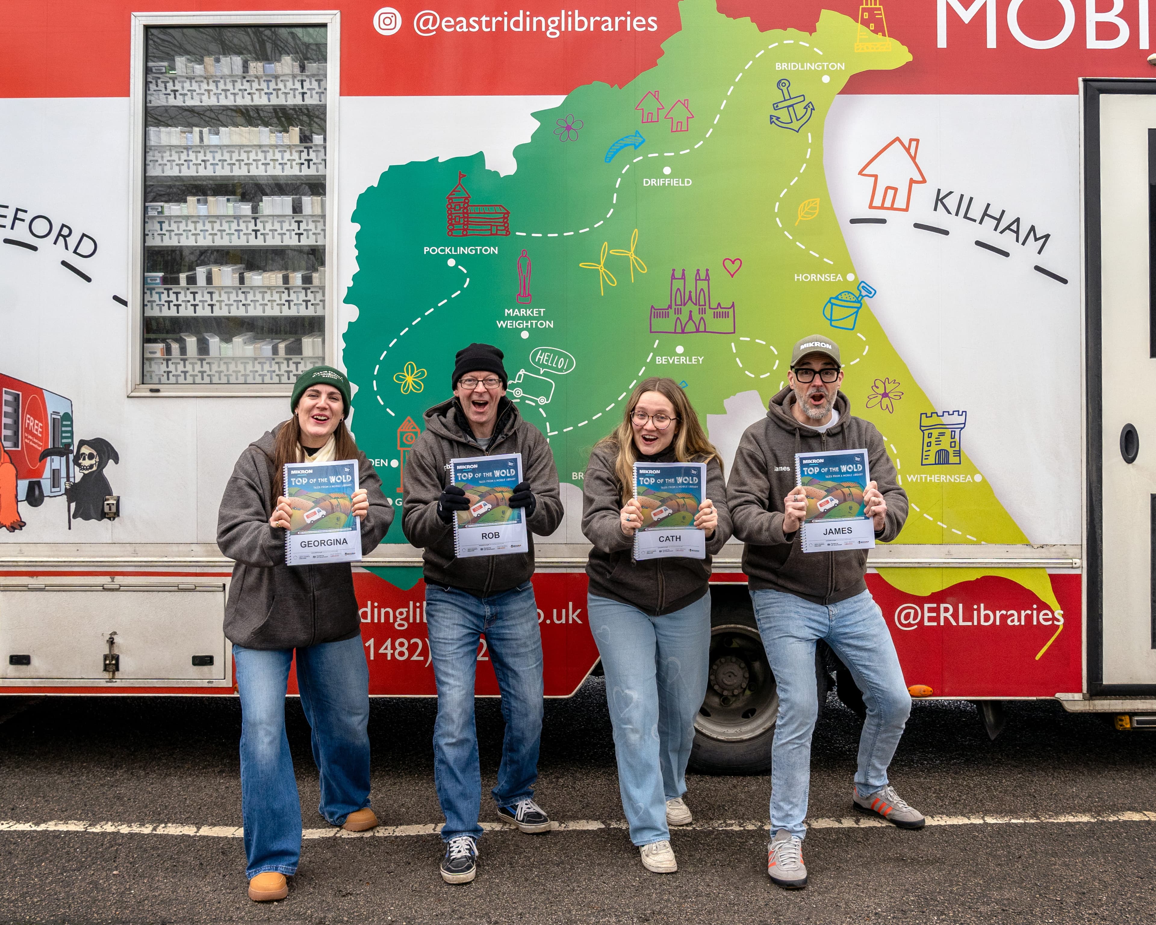 Four enthusiastic performers hold scripts in front of a colorful mobile library van, showcasing Yorkshire's map and landmarks.
