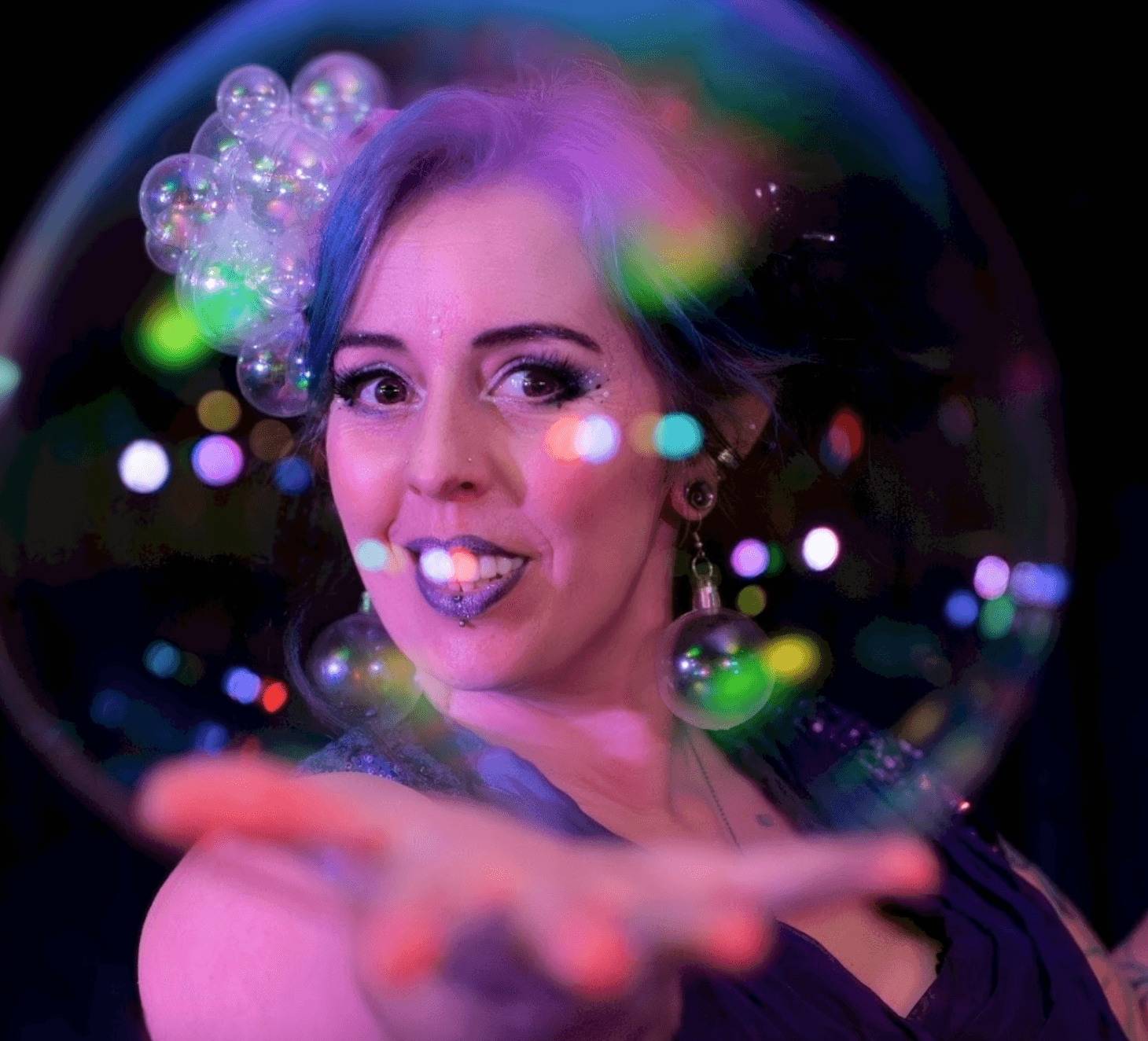A performer with colorful hair and sparkling makeup extends her hand, showcasing a large iridescent bubble against a vibrant backdrop.