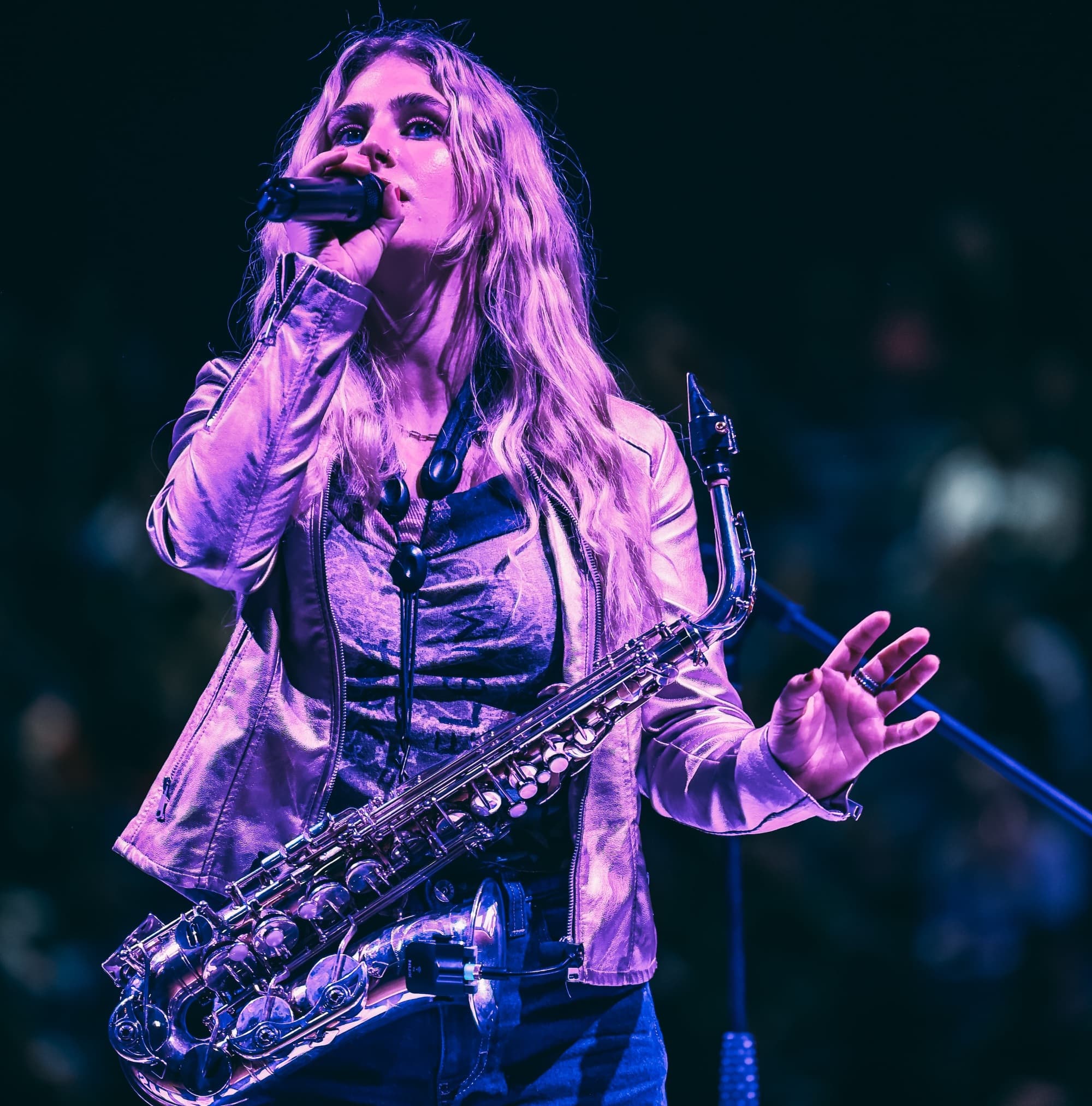 A female singer passionately performs with a saxophone, illuminated by stage lights, in a vibrant concert atmosphere.