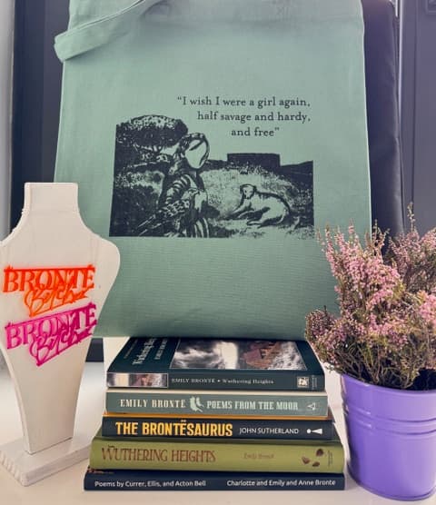 A green tote bag featuring a Brontë quote, surrounded by classic books and a purple pot of flowers.