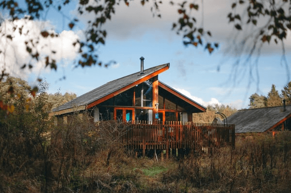A cozy wooden cabin nestled in a forest, surrounded by greenery and under a blue sky with fluffy clouds.