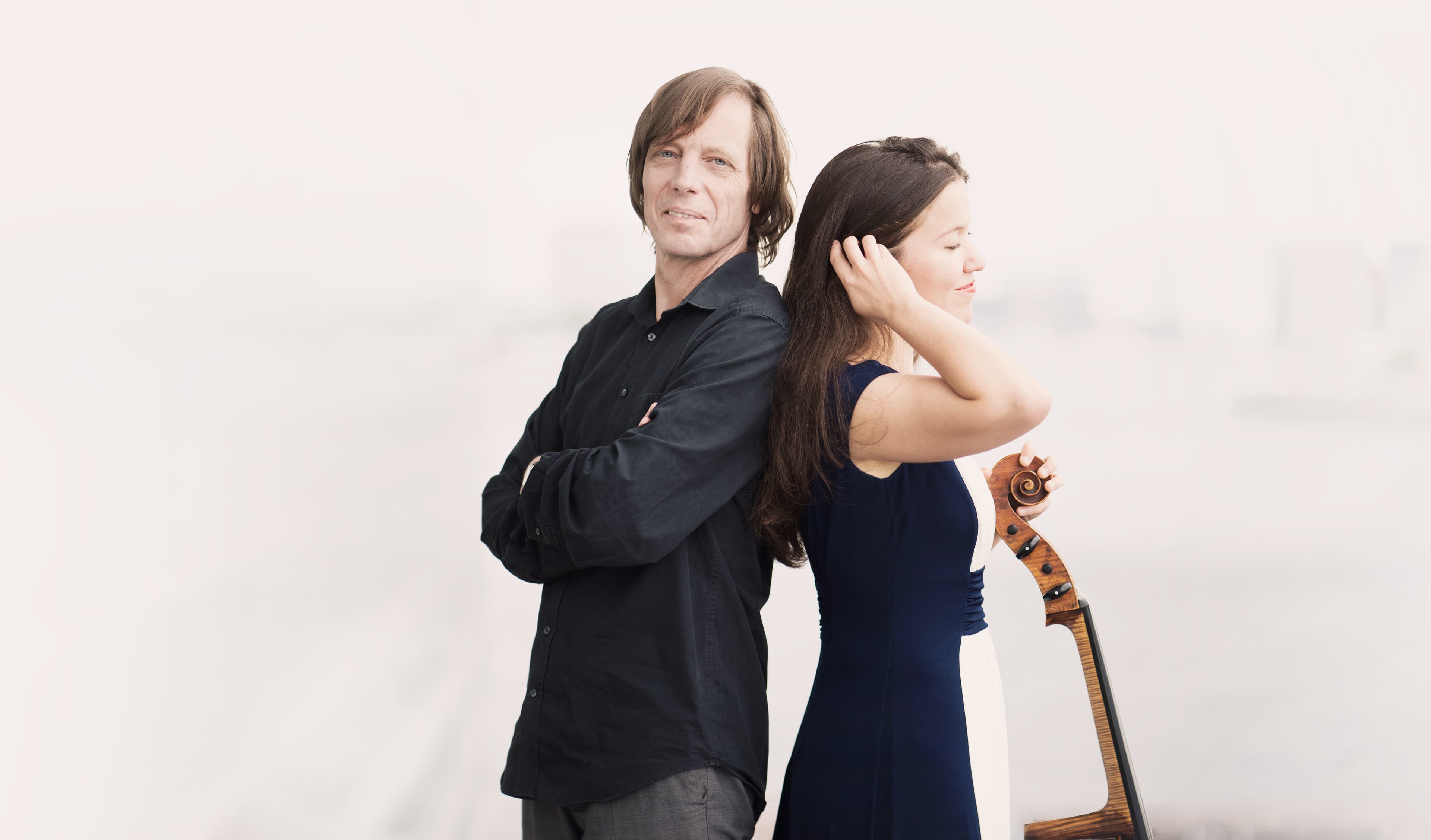 Two musicians pose back-to-back, one holding a violin, set against a soft, blurred background.