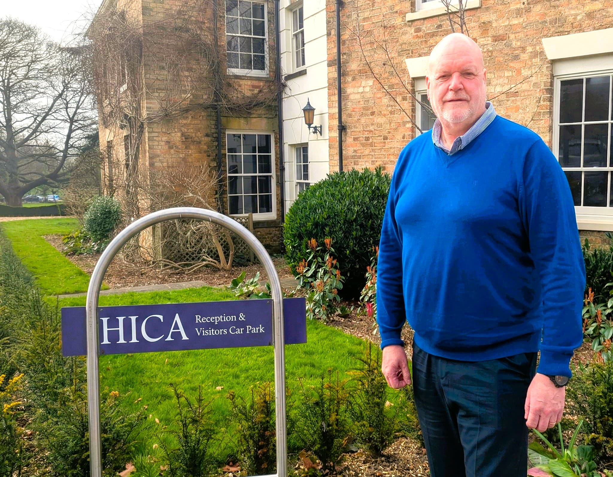 A man stands next to a sign for HICA, with a brick building and landscaped garden in the background.
