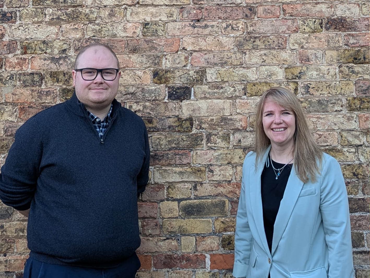 Two compliance support officers stand smiling against a textured brick wall, showcasing a professional and welcoming atmosphere.