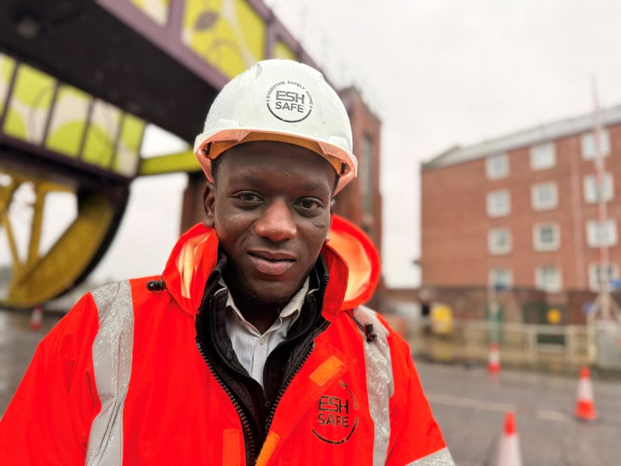 Apprentice wearing a reflective orange jacket and hard hat stands near Hull’s Drypool Bridge, with buildings in the background.
