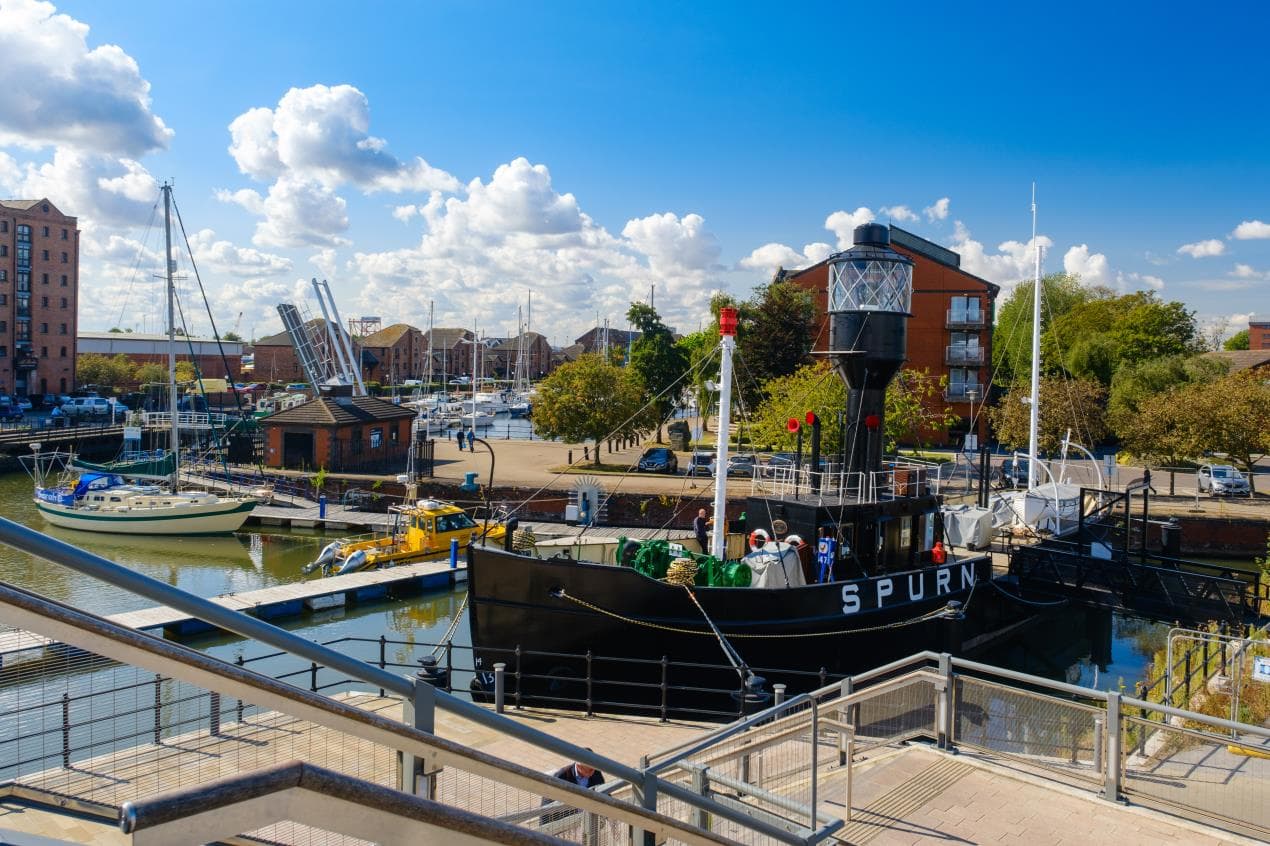 Spurn Lightship moored at Hull’s marina, surrounded by boats, historic buildings, and vibrant greenery under a blue sky.