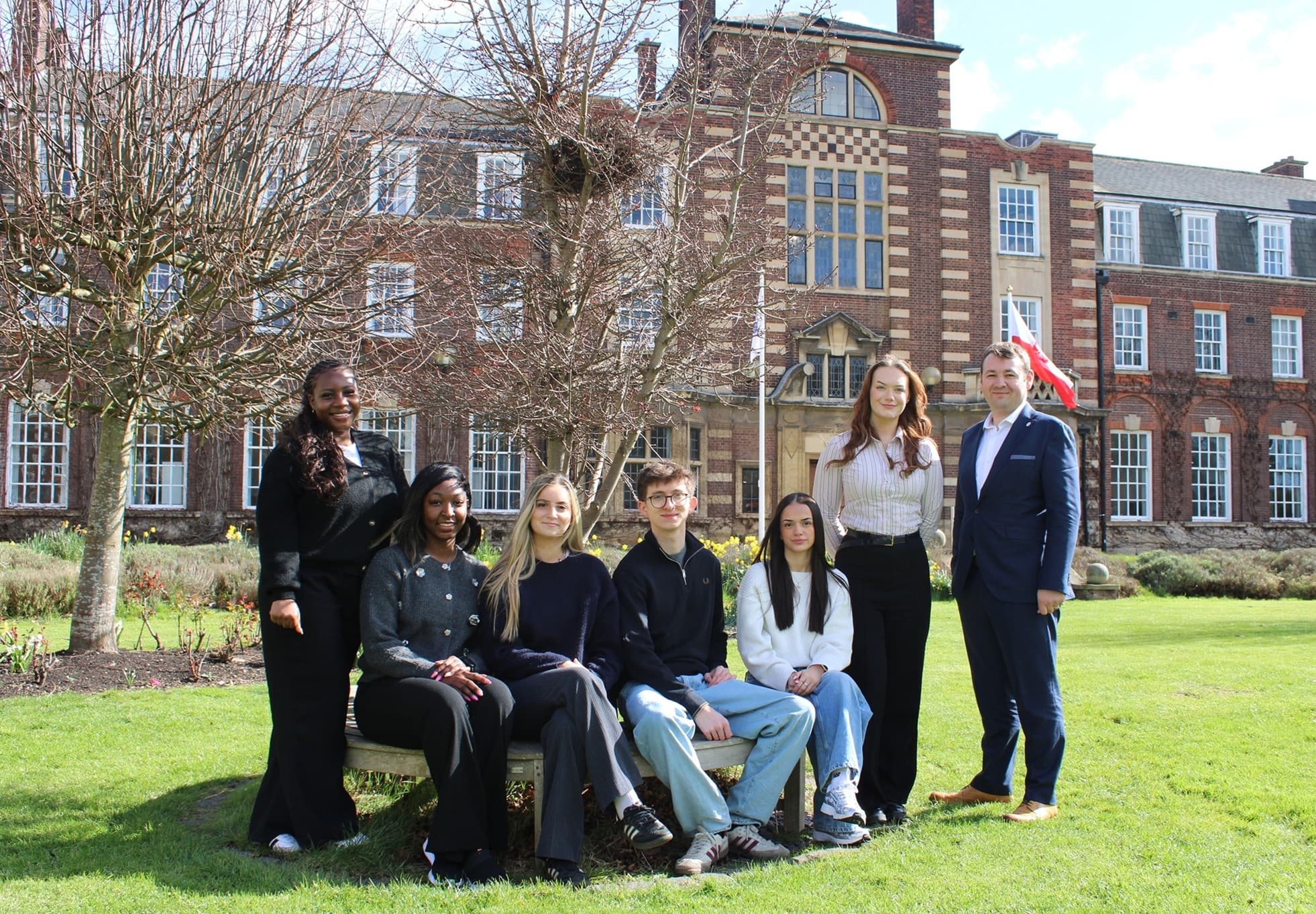 Students and faculty pose on a grassy lawn in front of a historic brick building, under a clear blue sky.