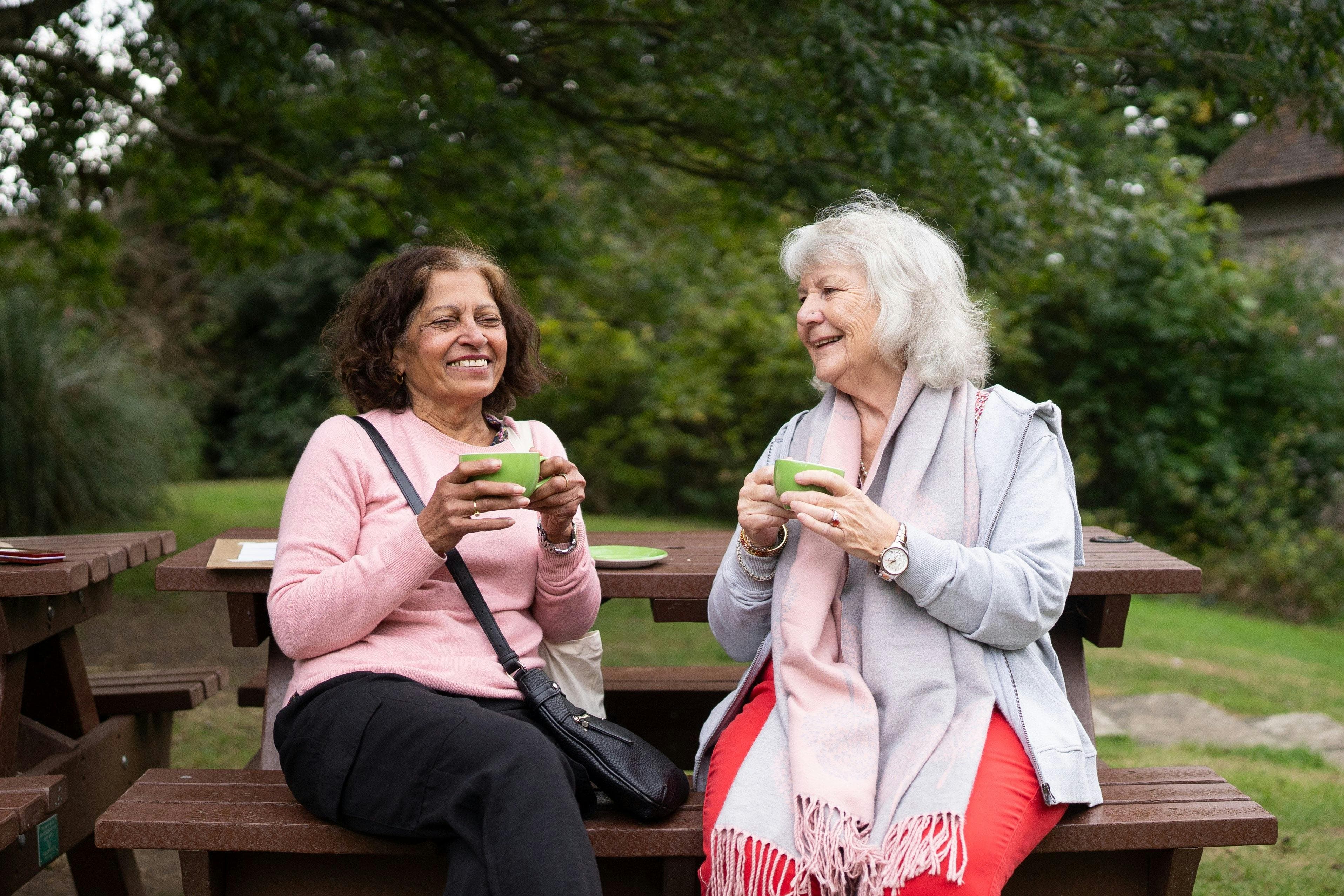 Two older women sit at a picnic table, enjoying tea and laughing together in a lush green garden setting.