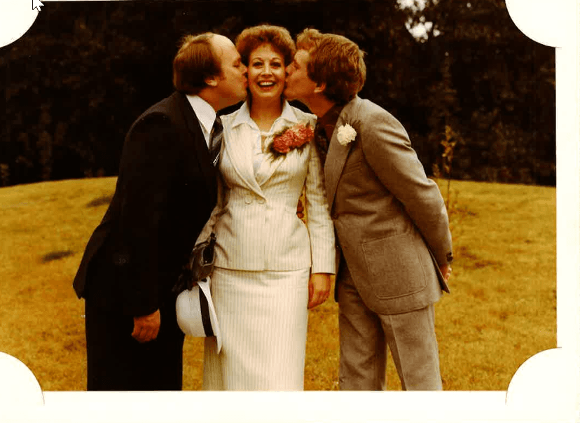 Three smiling individuals in formal attire, with one woman in a white suit, surrounded by greenery in a warm, celebratory atmosphere.