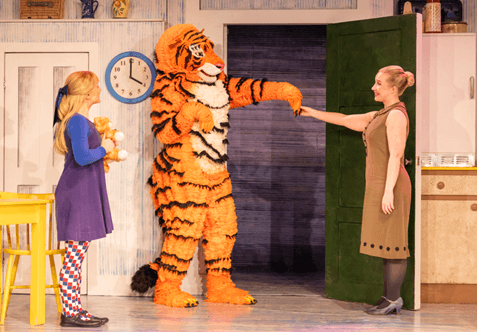 A vibrant stage scene featuring a tiger character greeting a woman, with a child holding a toy, set in a colorful kitchen.