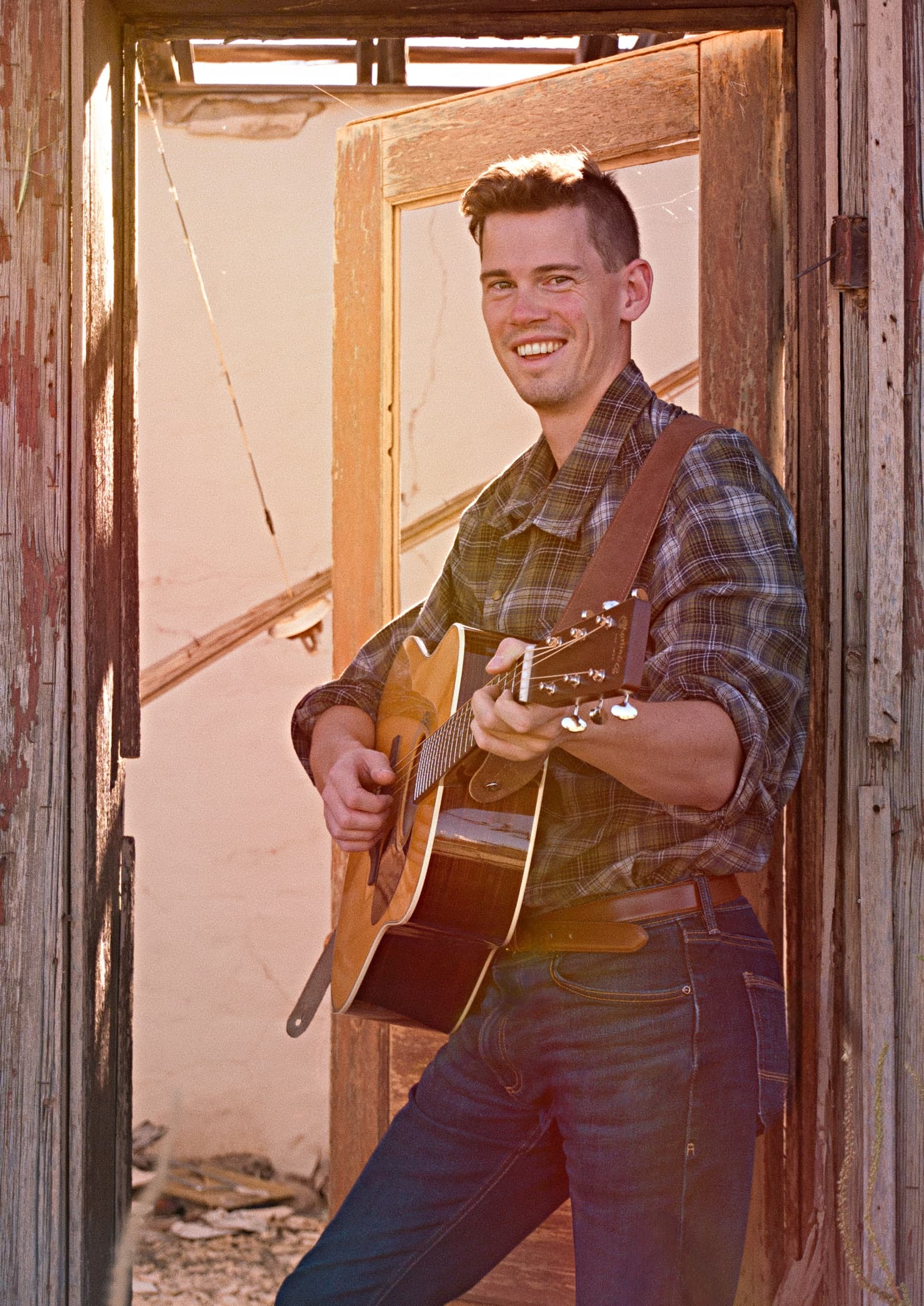 A young man in a plaid shirt and jeans smiles while holding a guitar, standing in a rustic doorway with warm sunlight.