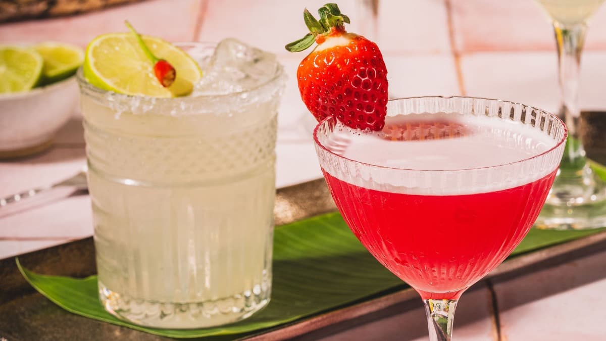 Two colorful cocktails sit on a dark tray, garnished with lime and strawberry, against a vibrant, tropical backdrop.