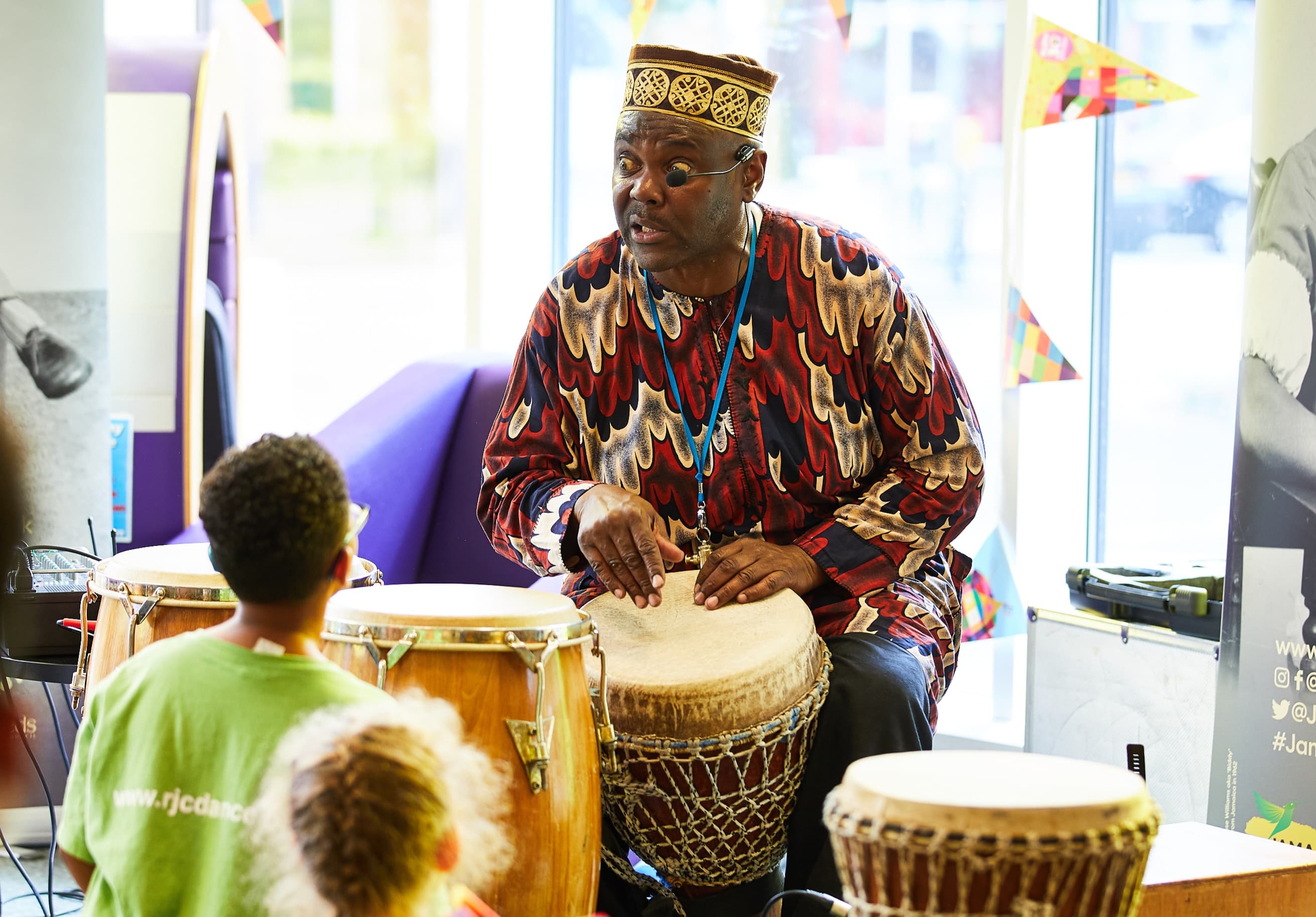 A drummer in colorful traditional attire engages children with percussion instruments at a lively event in Huddersfield.