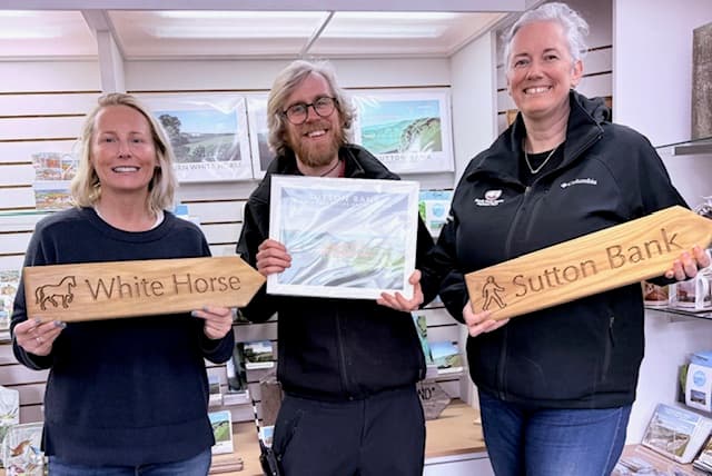 Three smiling individuals hold wooden directional signs for Sutton Bank and the White Horse, showcasing local tourism.
