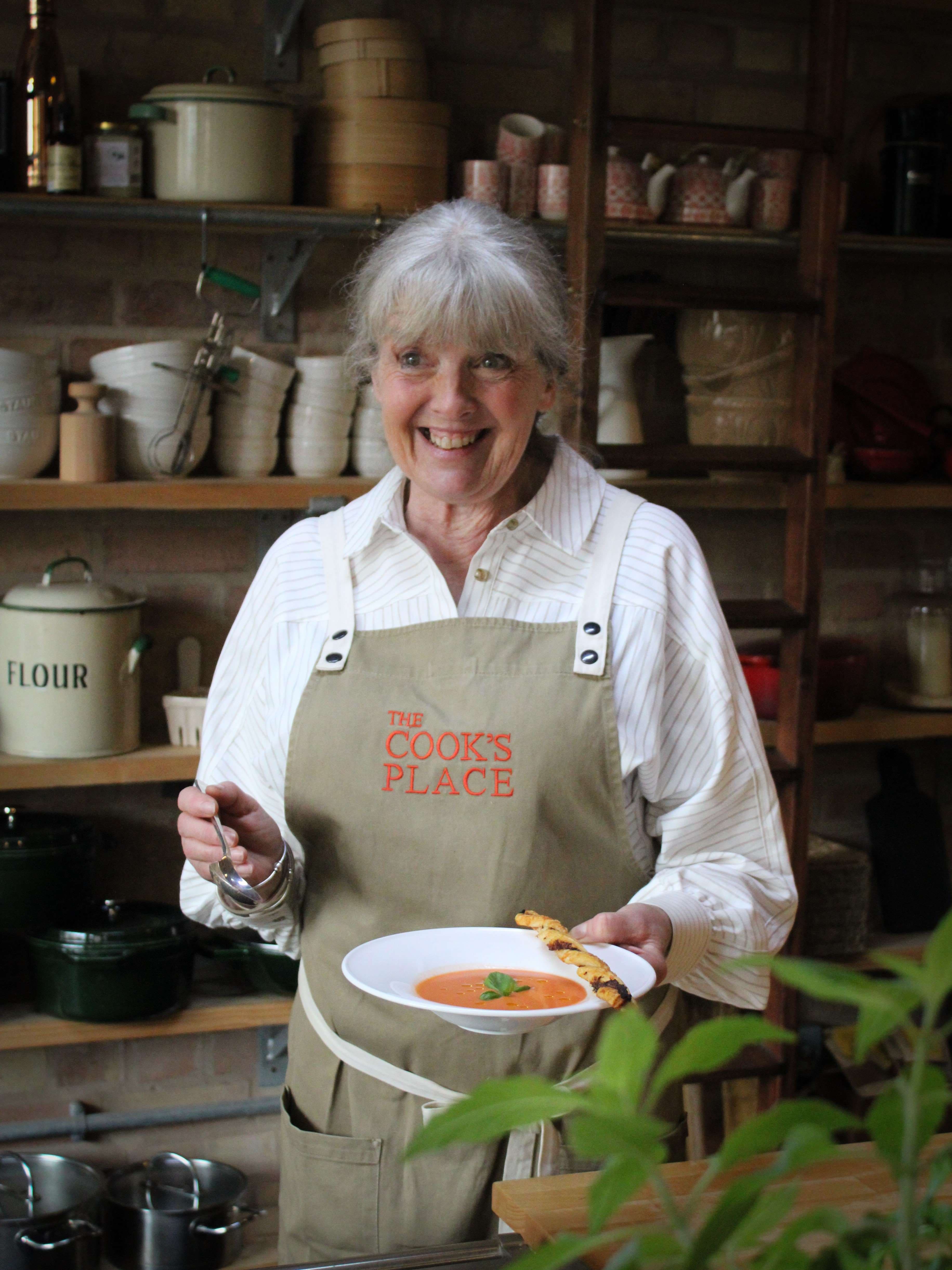 A smiling chef in an apron holds a bowl of tomato soup garnished with basil, surrounded by rustic kitchenware.