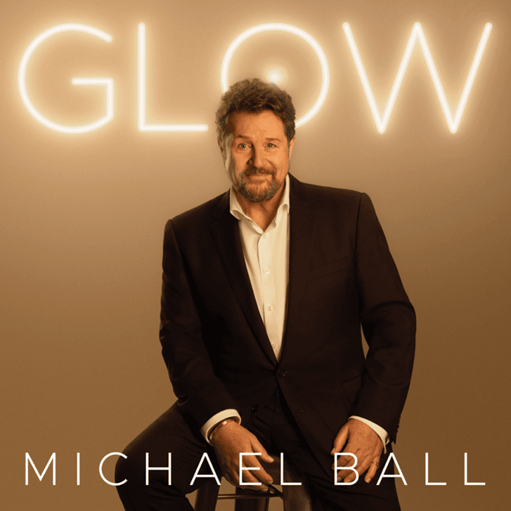 Michael Ball sits confidently on a stool, dressed in a smart suit, with a warm glow from the backdrop illuminating the scene.