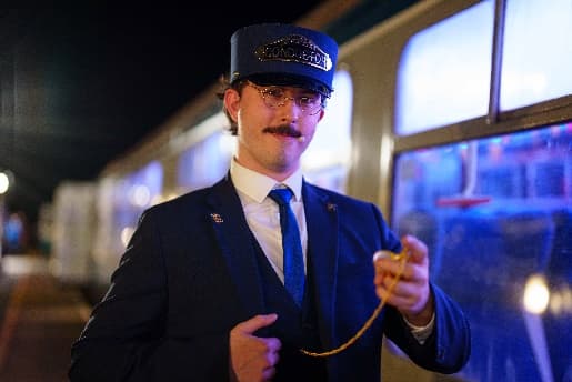 Conductor in a blue uniform and hat stands by a lit train carriage at Wensleydale Railway, creating a festive atmosphere.