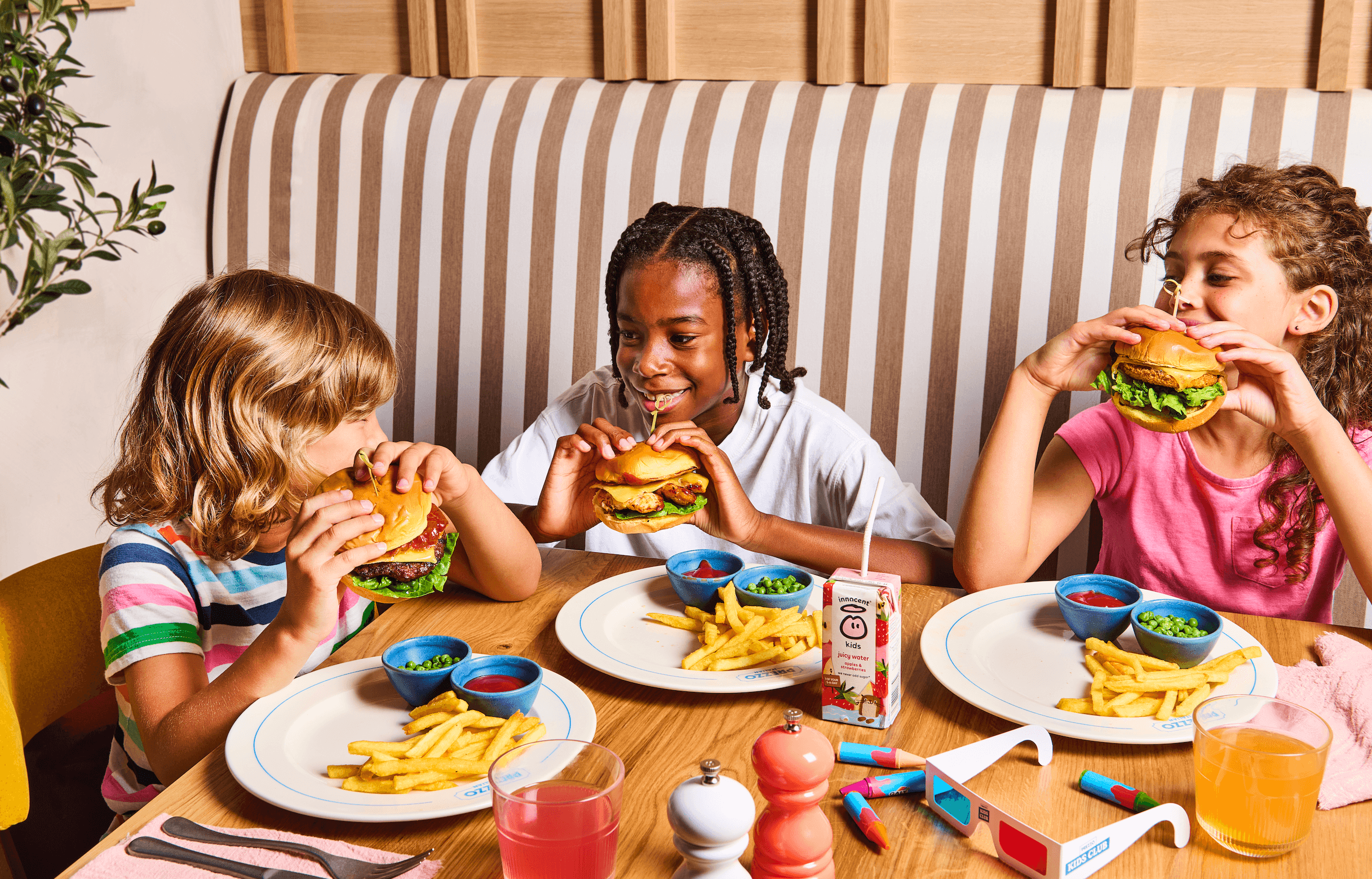 Three children enjoy burgers and fries at a table, smiling and sharing a fun moment in a cozy restaurant setting.
