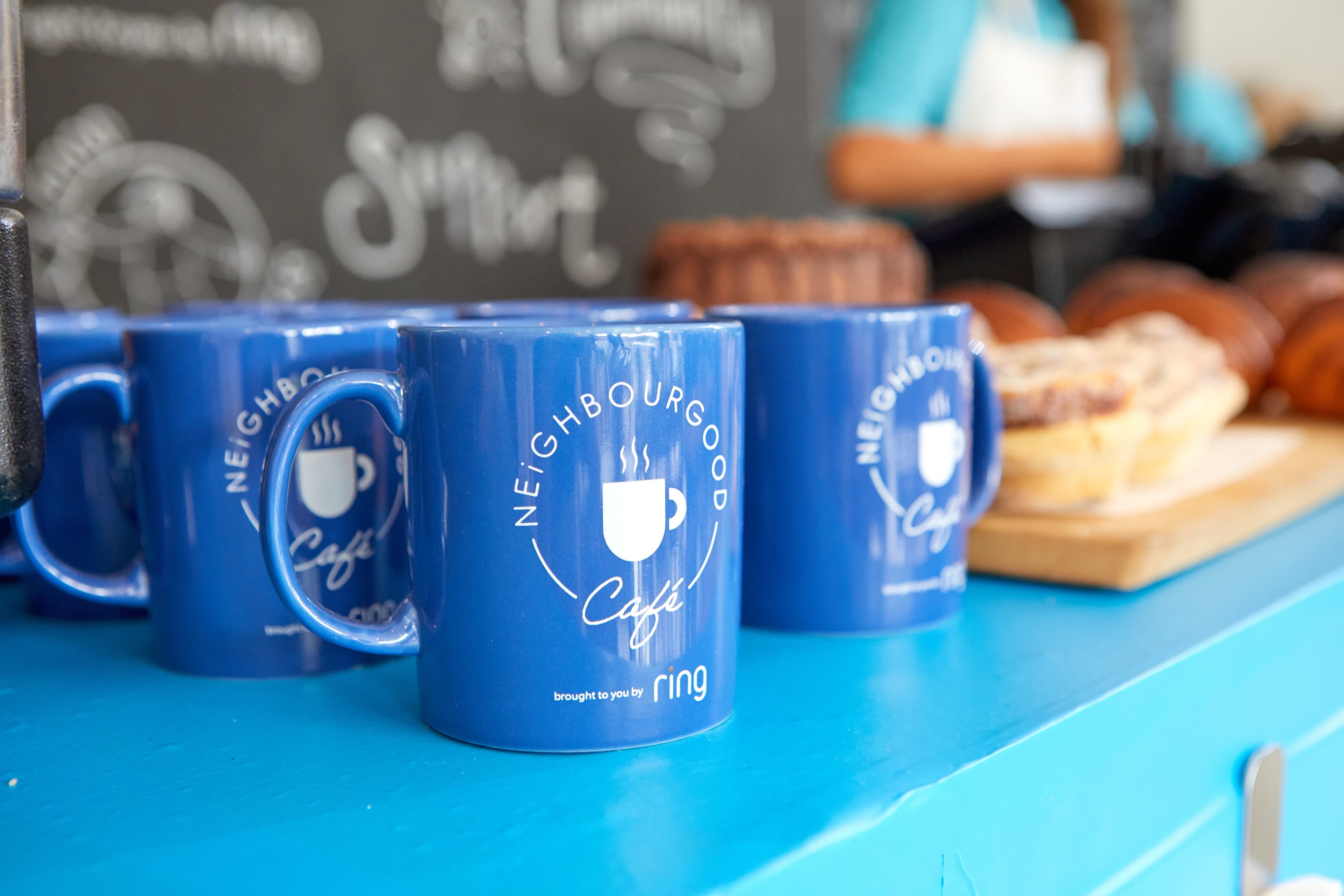 Bright blue mugs with "NeighbourGood Café" logo sit on a vibrant counter, with baked goods and a chalkboard backdrop.