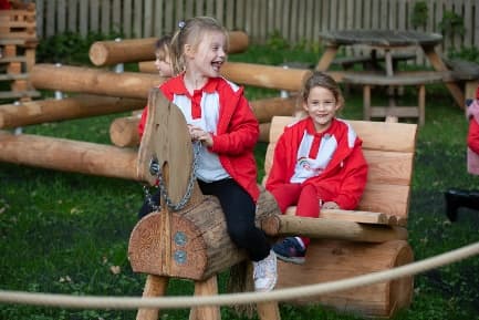 Children in red jackets joyfully play on wooden horse and cart structures in a green outdoor area at Abbeydale Industrial Hamlet.
