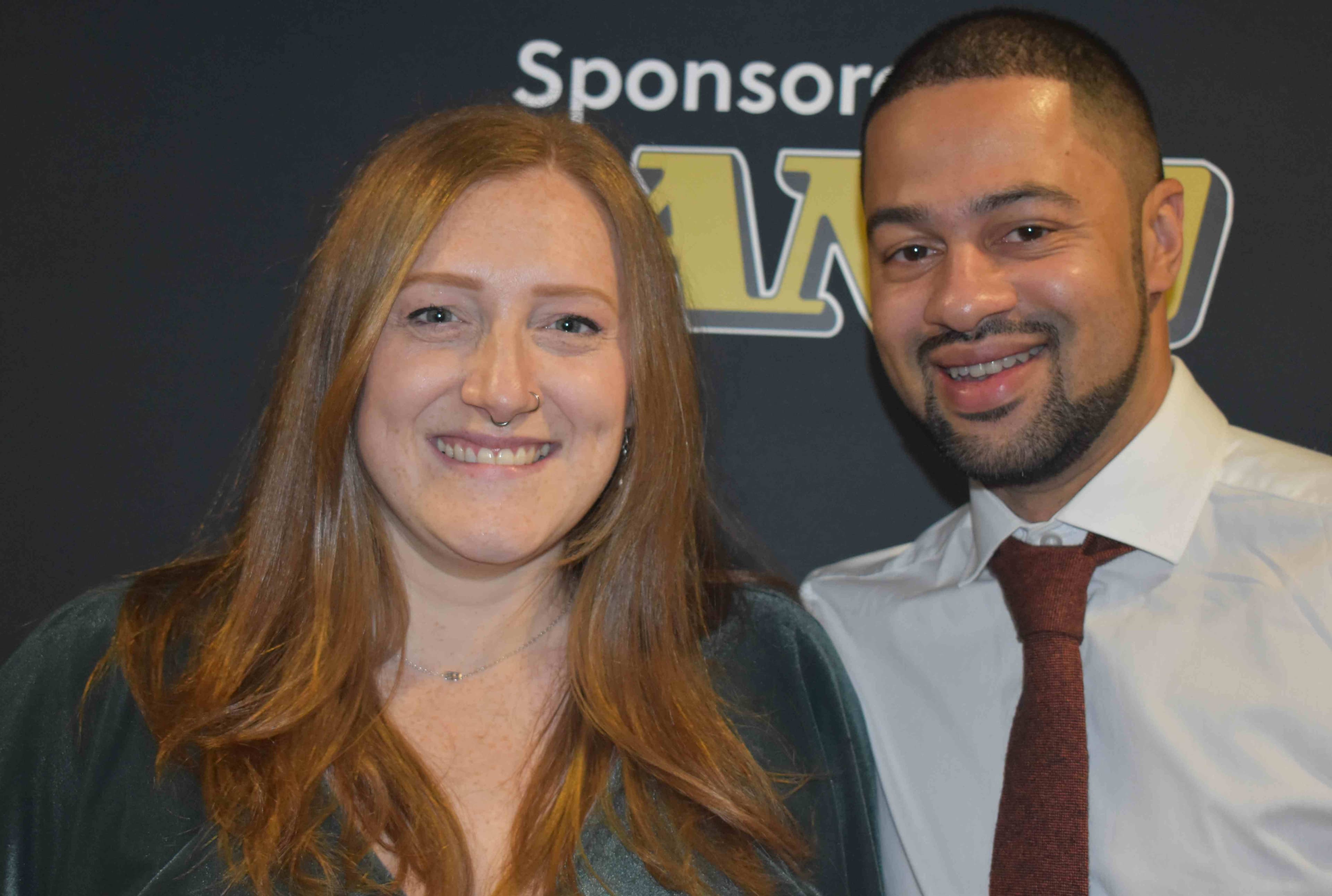 Smiling woman with long red hair in a green dress stands beside a man in a white shirt and maroon tie, celebrating an award.