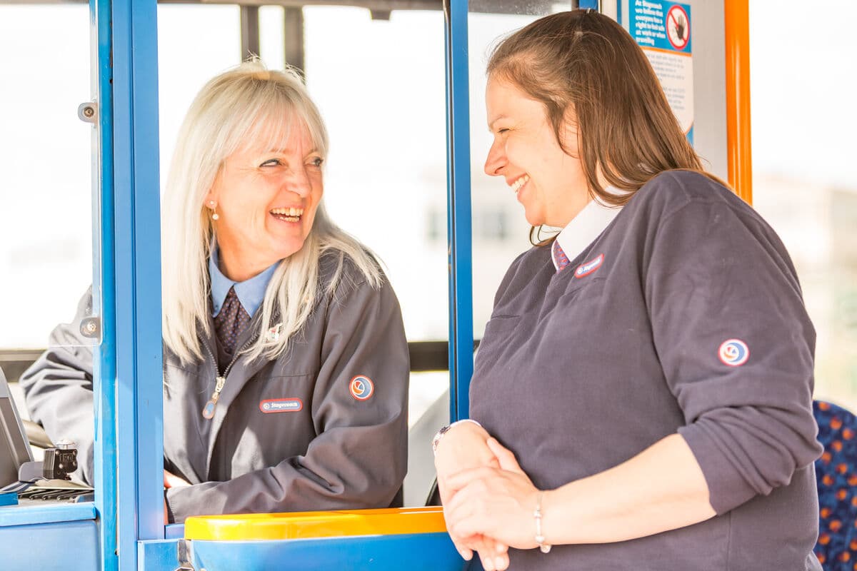 Two smiling bus operators in uniforms chat at a bus station, with vibrant blue and orange surroundings.