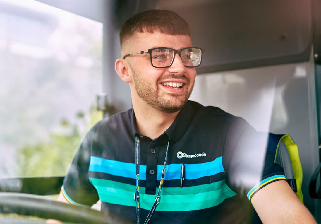 A smiling Stagecoach driver wearing glasses, seated in a bus, with green foliage visible outside the window.