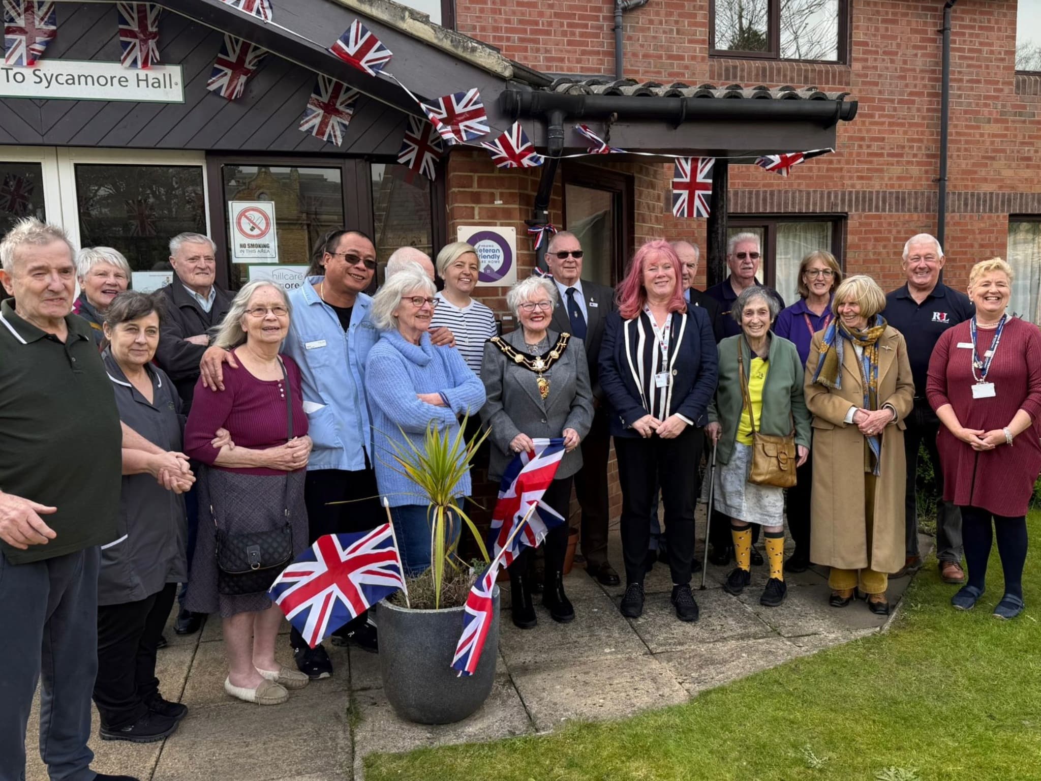 A diverse group of people gathered outside Sycamore Hall in Ripon, adorned with Union Jack flags, celebrating together.