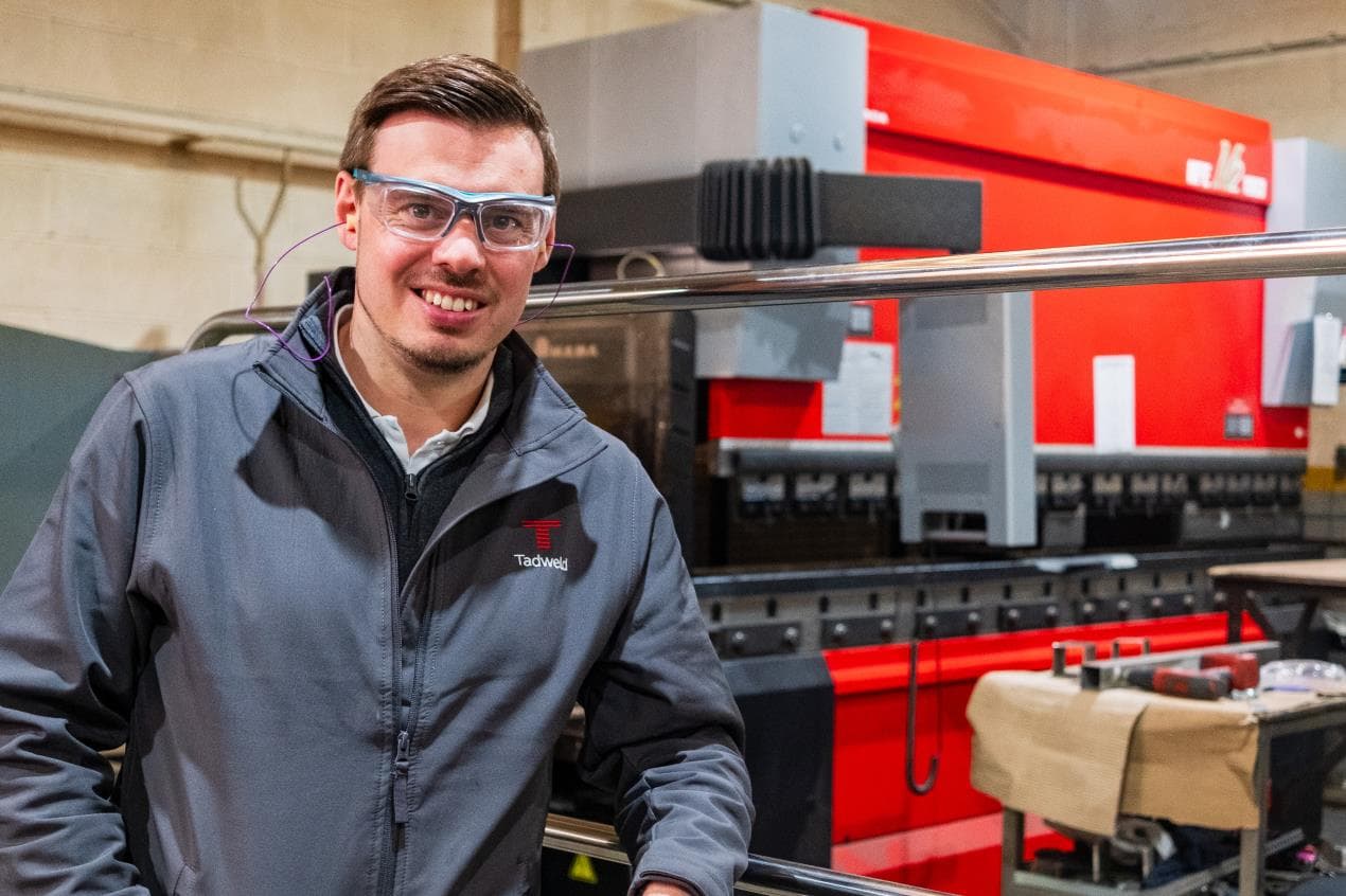 A man in safety glasses and a Tadweld jacket stands in a manufacturing facility, with machinery in the background.