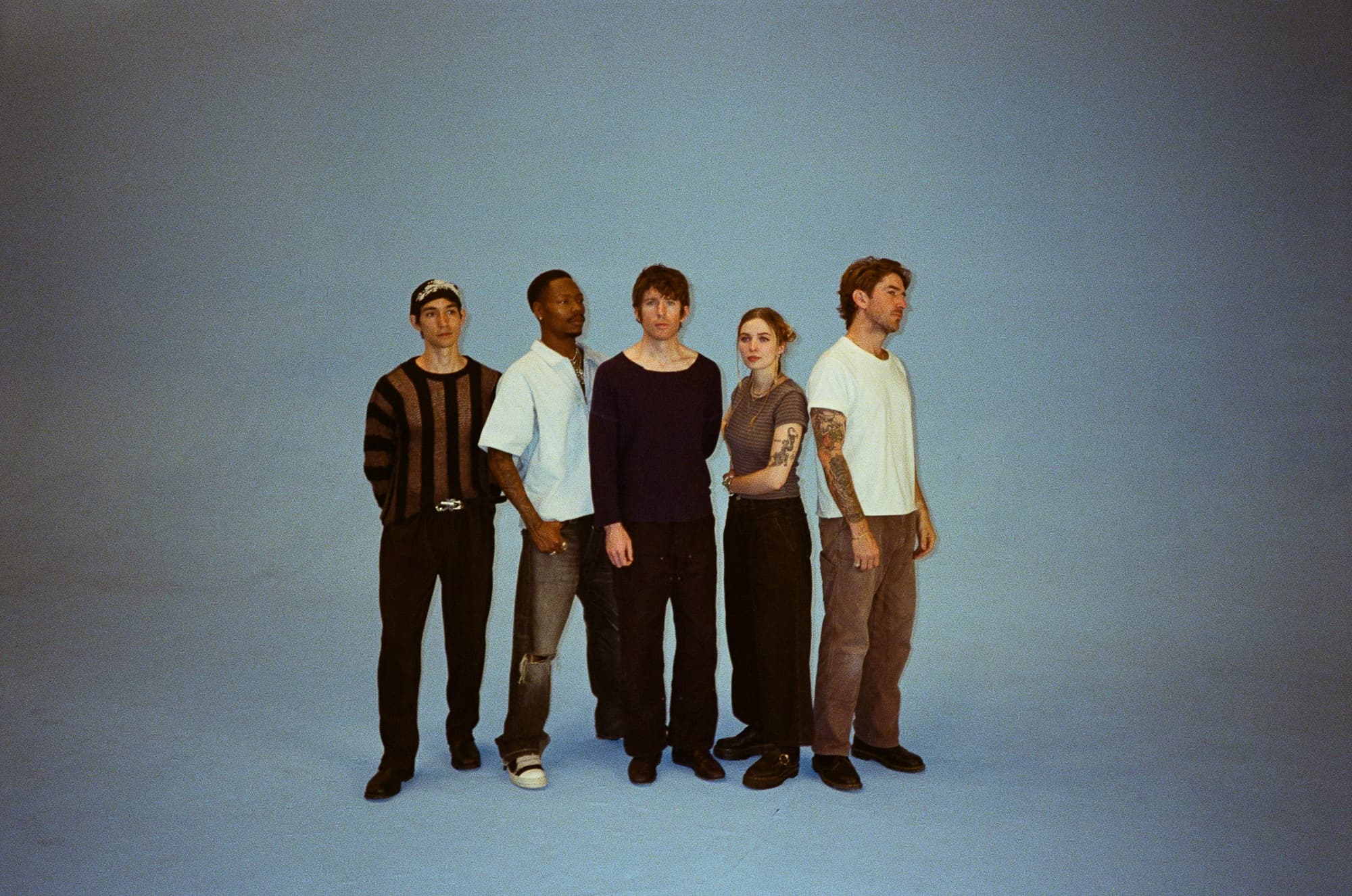 Five band members stand together against a plain blue backdrop, each wearing casual clothing with varied styles and expressions.