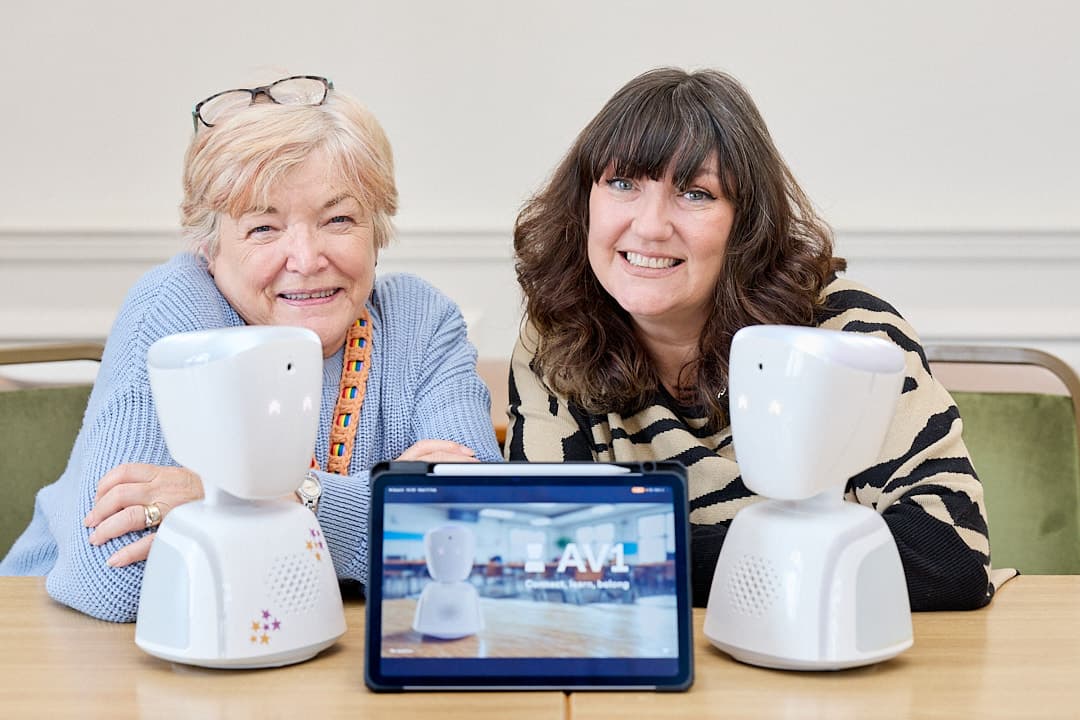 Two women smile at the camera, flanking classroom robots and a tablet, in a bright, inviting educational setting.