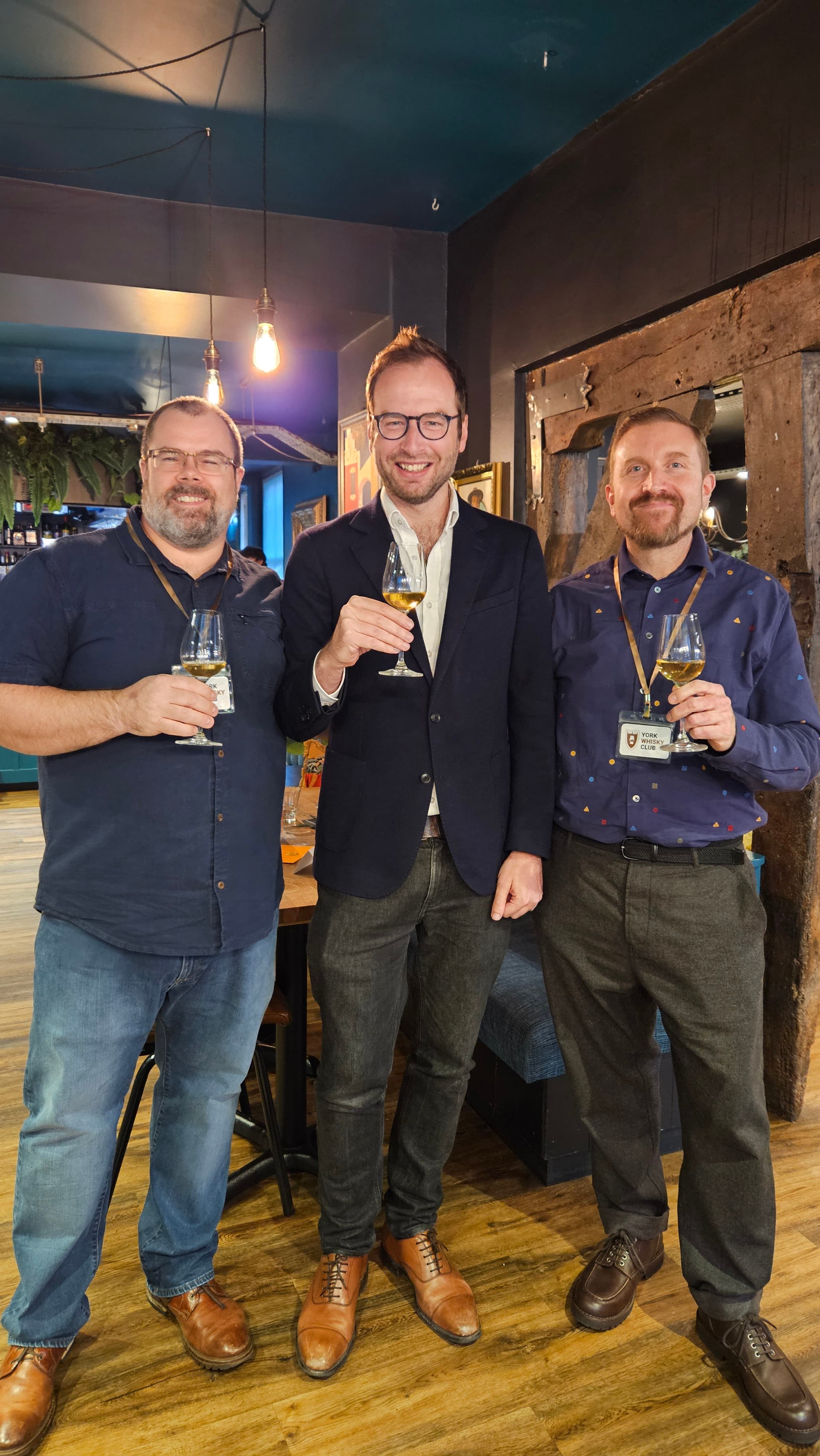 Three men in smart casual attire hold drinks, smiling in a cozy, stylish bar setting with warm lighting and rustic decor.