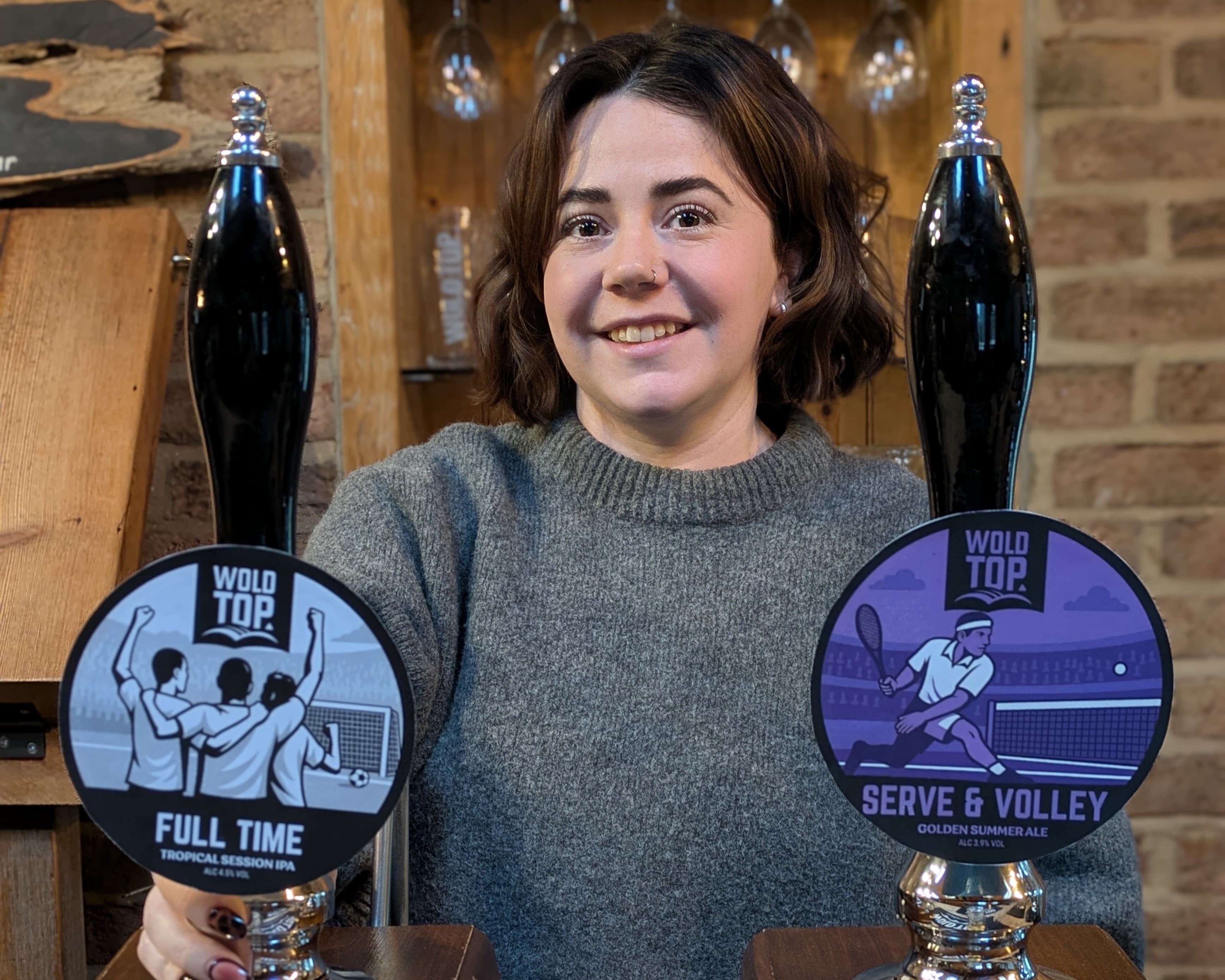 A woman smiles behind two beer taps labeled "FULL TIME" and "SERVE & VOLLEY," set against a rustic brick backdrop.