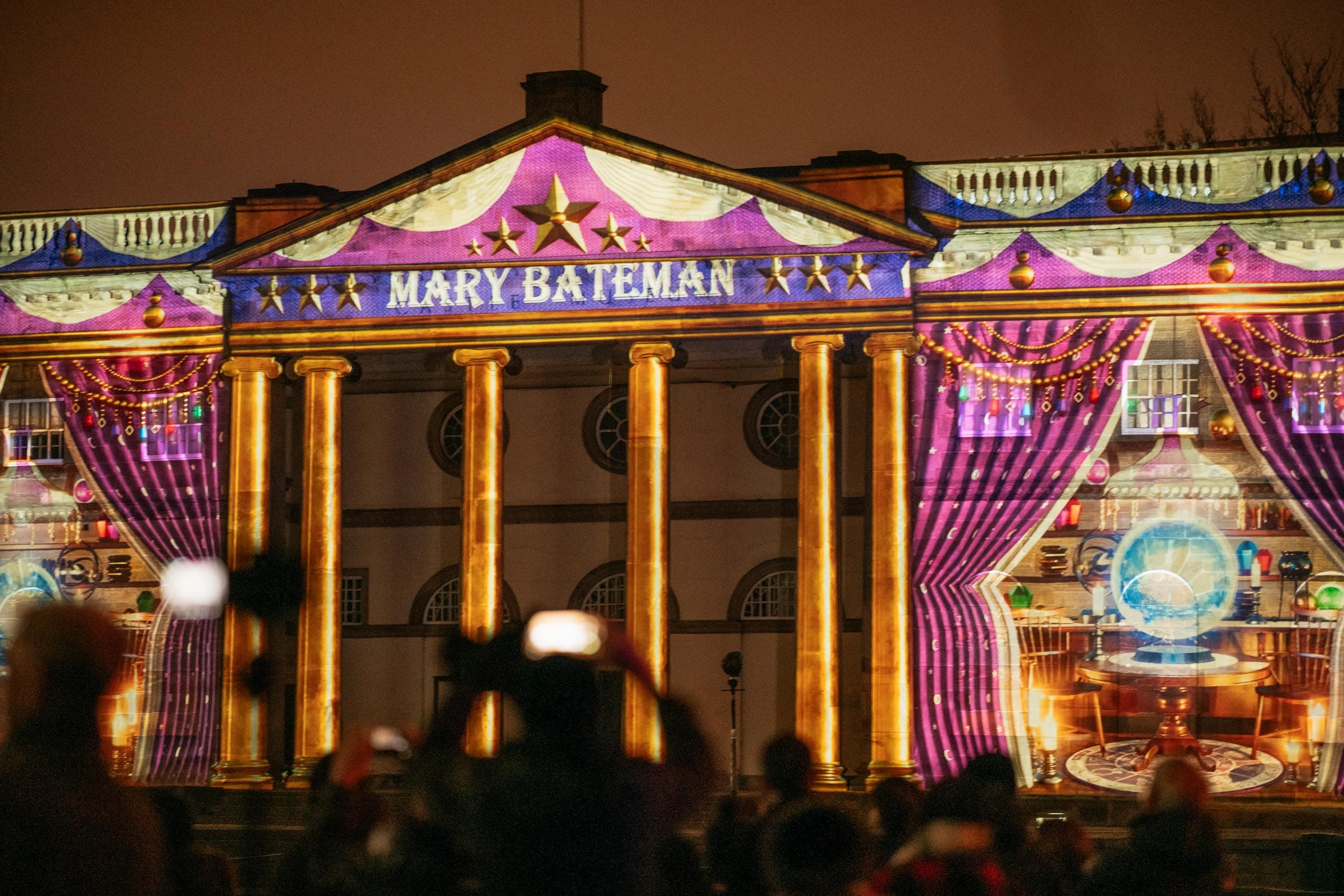 Vibrant light projections illuminate the grand façade of a historic building, with a crowd admiring the spectacle at night.