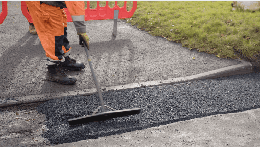 A worker in orange overalls smooths freshly laid asphalt on a road, with traffic barriers and green grass nearby.