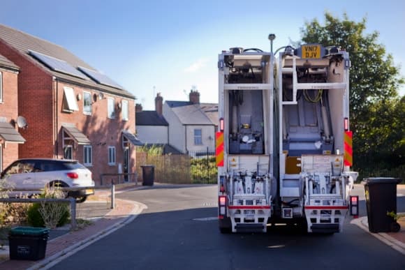 Wheeled recycling bins line a residential street in York, with a refuse truck collecting waste under a clear blue sky.