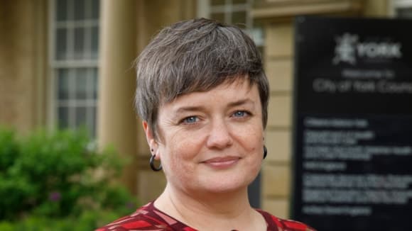 A woman with short hair stands in front of the City of York Council building, smiling, with greenery in the background.