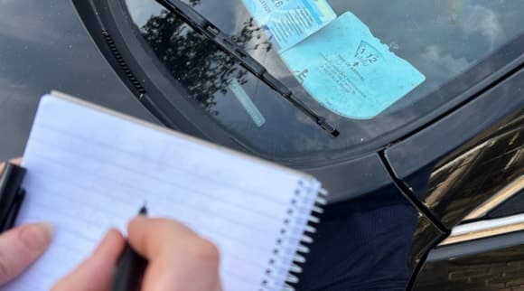 A close-up of a hand writing in a notebook next to a car displaying a blue disabled parking badge on its windshield.