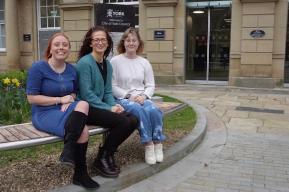 Three women sit on a bench outside the City of York Council building, smiling amidst blooming daffodils and stone pathways.
