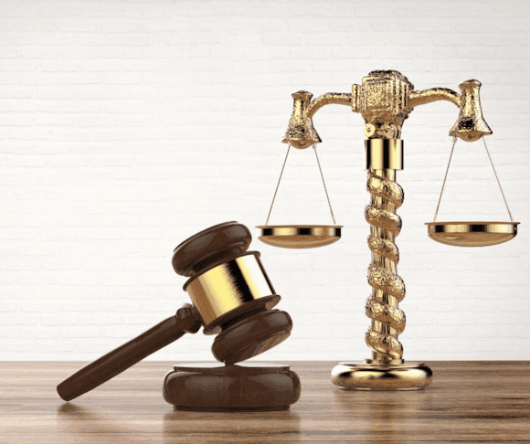 A wooden gavel rests beside a golden balance scale on a polished wooden surface, symbolizing justice and legal proceedings.