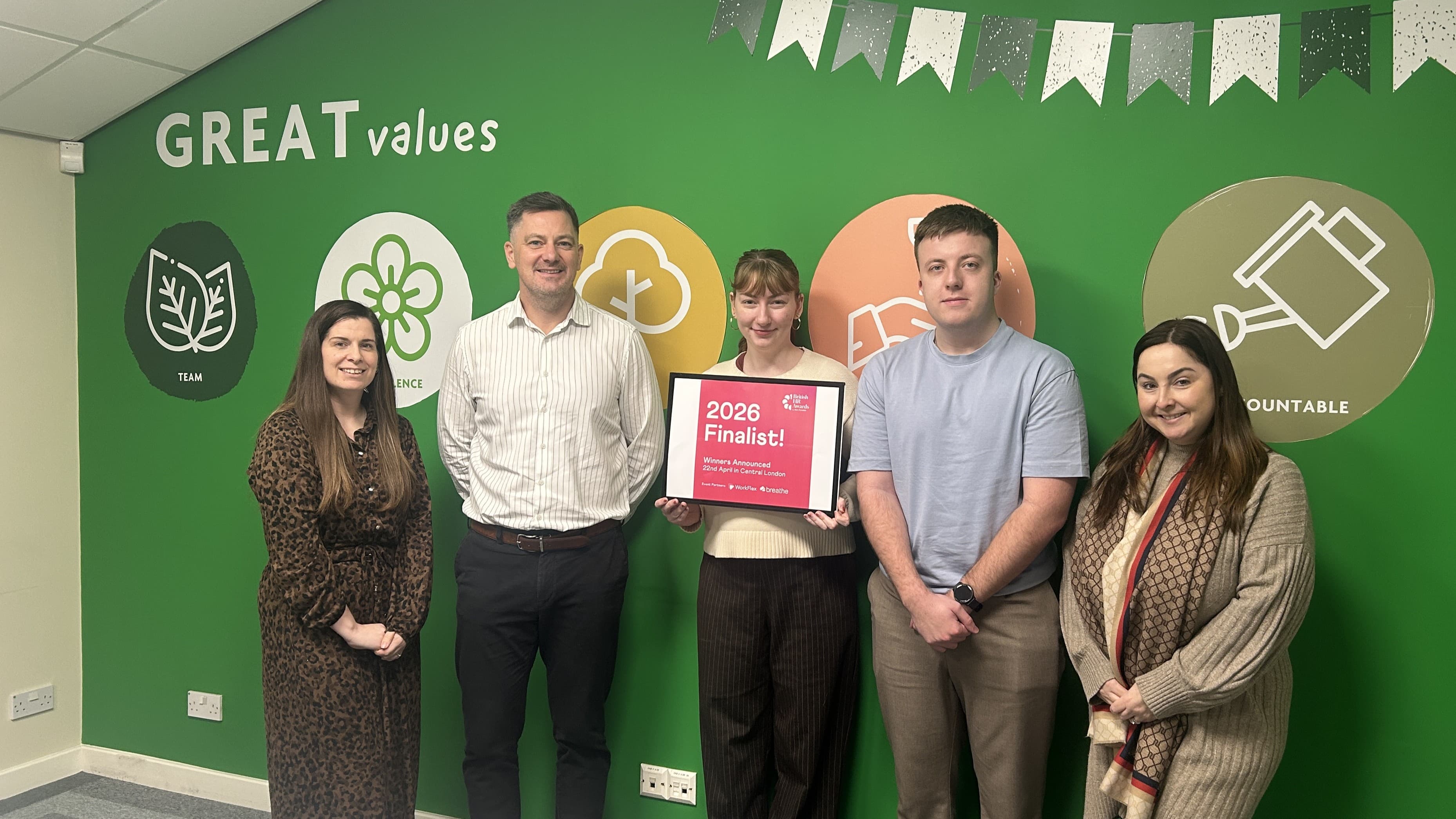 Five team members stand in front of a green wall displaying the words "GREAT values" and colorful circular icons, holding a finalist sign.