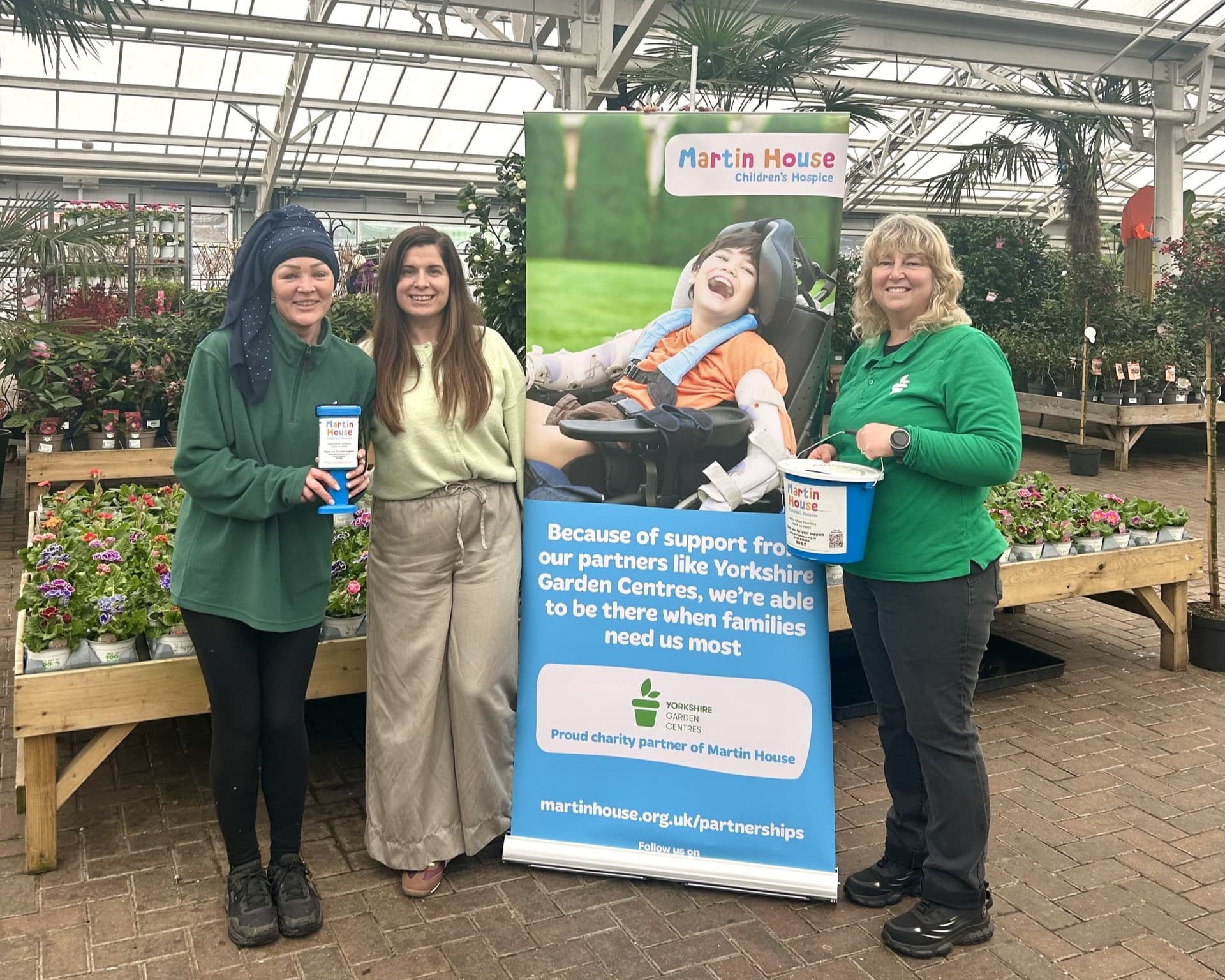 Three women hold collection buckets beside a banner for Martin House Children's Hospice in a vibrant garden centre.