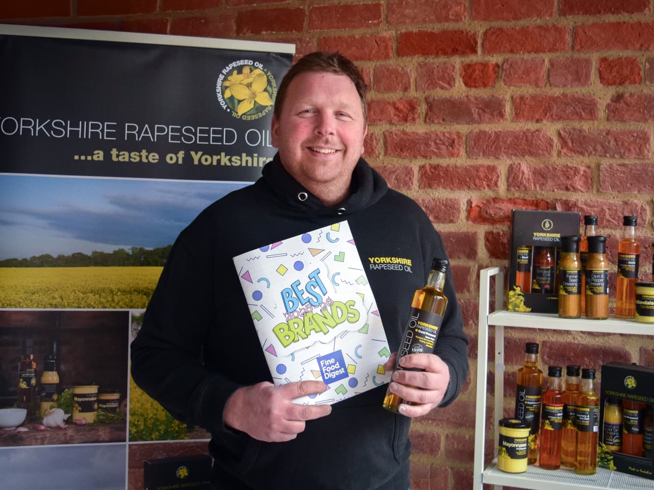 A man in a black hoodie holds a "Best Brands" award beside shelves of rapeseed oil products against a brick wall backdrop.