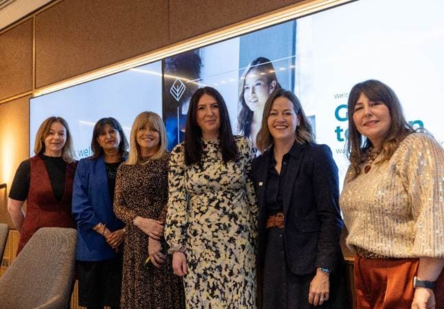 Six women stand together in a modern conference room, smiling and dressed in professional attire, with a large screen behind them.