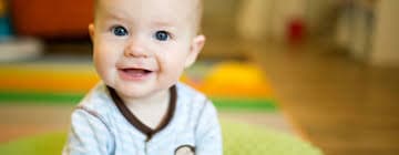 Smiling baby in blue pajamas sitting on a colorful rug