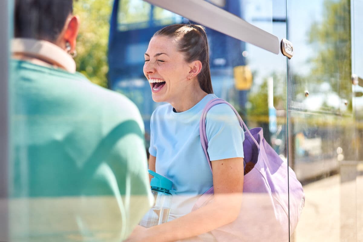 Woman laughing at a sunny bus stop with a backpack