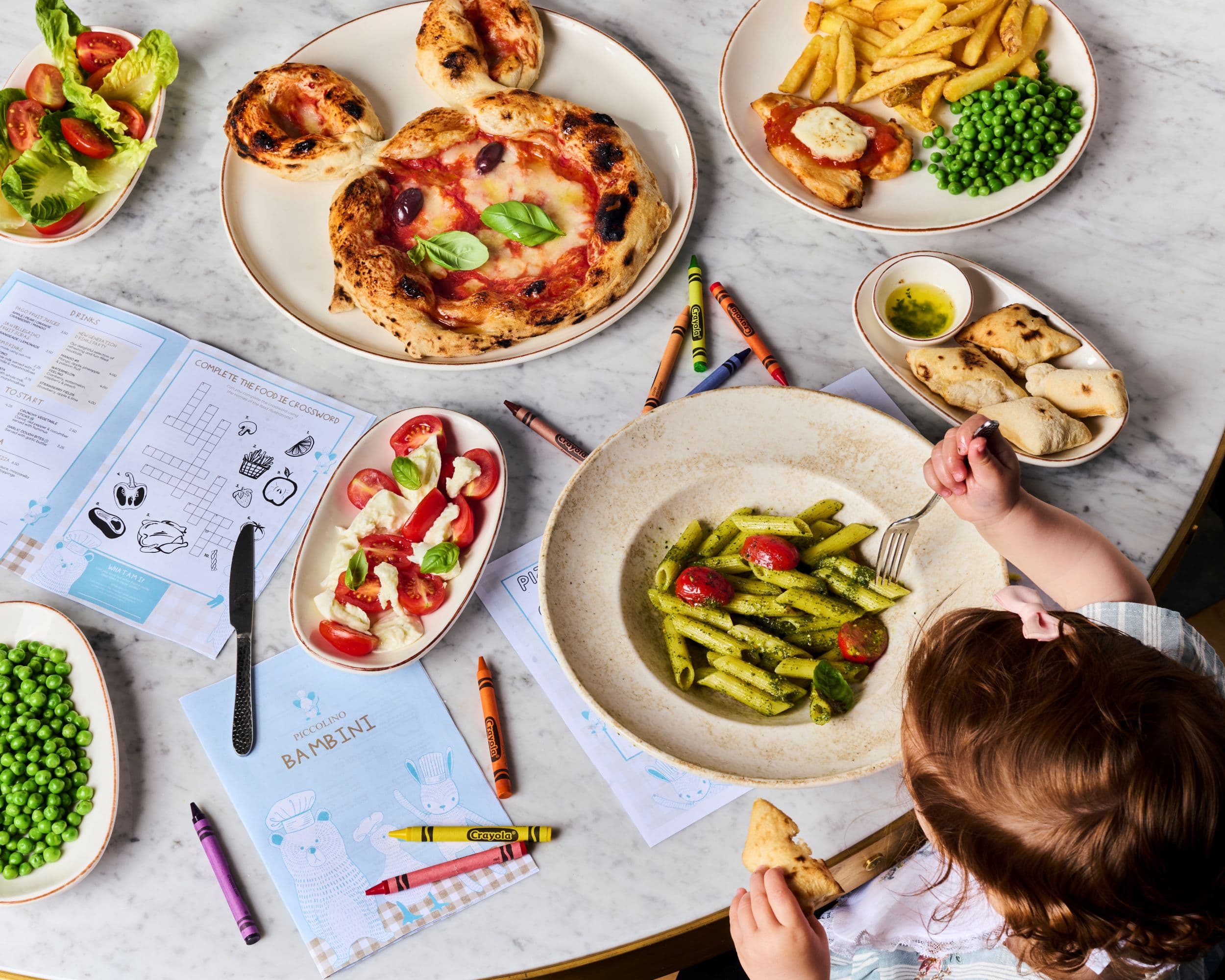 Child enjoying pasta at a table with pizza and coloring sheets