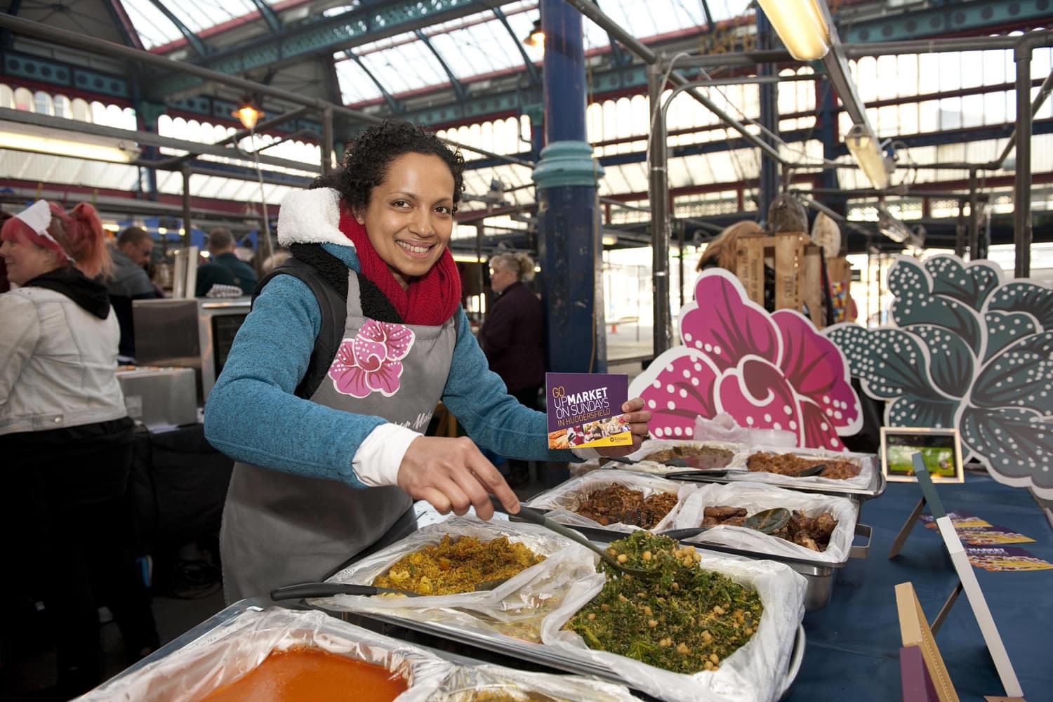 Vendor serving diverse dishes at a bustling indoor market with decorative stalls and overhead glass panels.